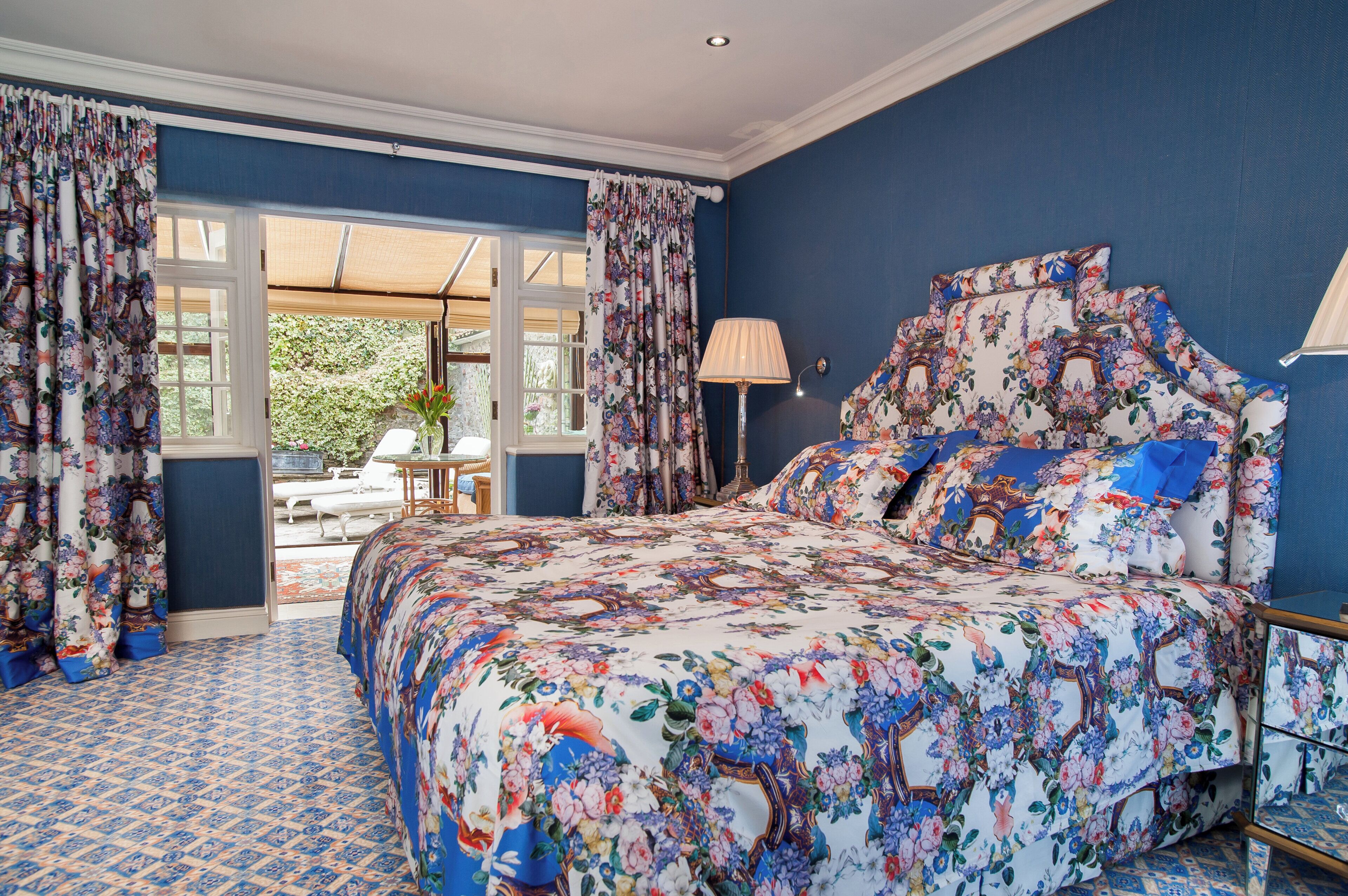 A bedroom with a floral bedspread, a large window with curtains, and a view of the outdoors.