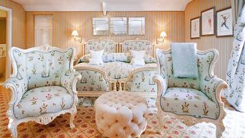 A room with a bed, two armchairs, a tufted ottoman, and a patterned carpet.