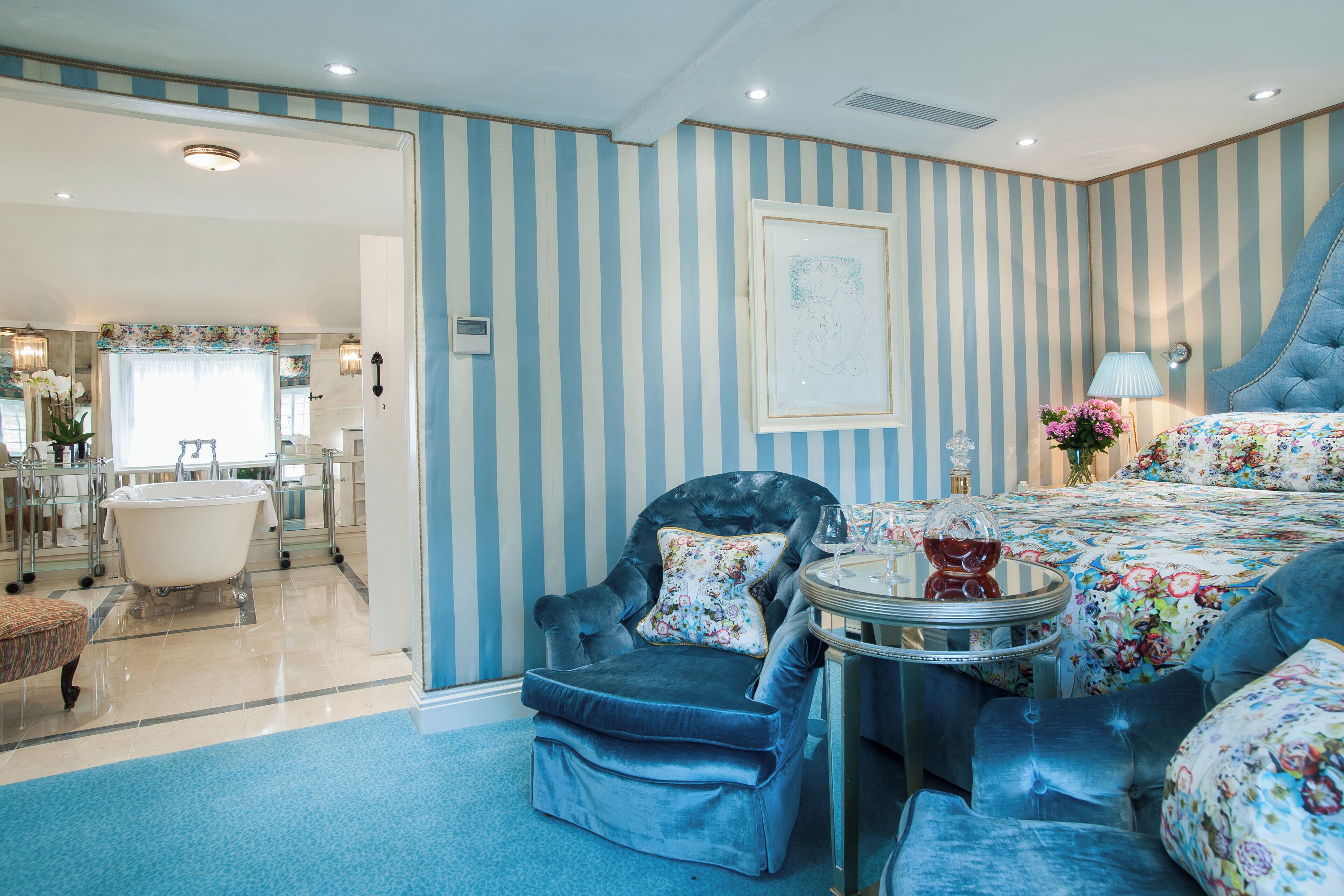 A room with a striped wall, a bed with floral bedding, a blue armchair, a round table with a vase, and a bathtub in the background.