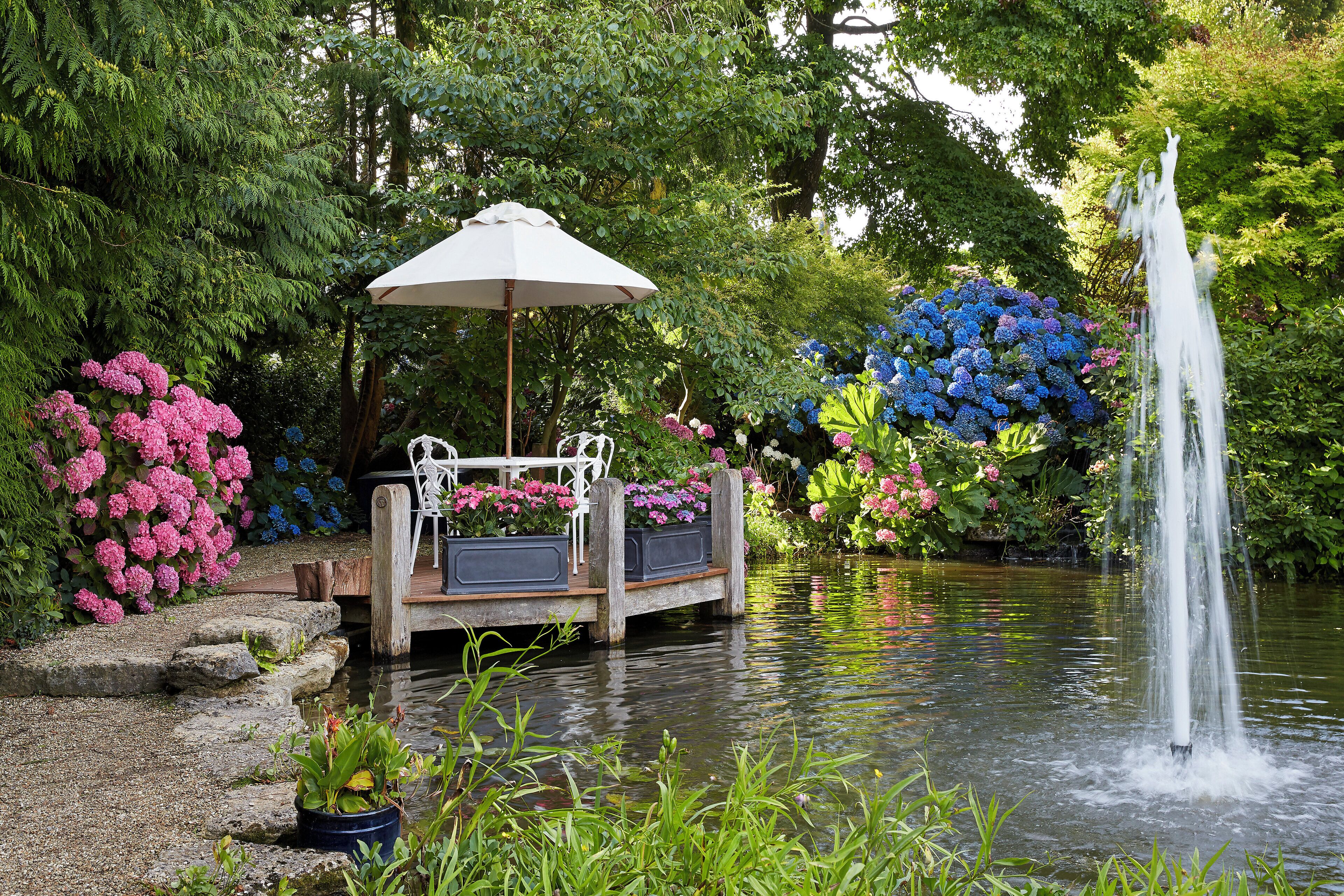 A serene garden with a pond, a wooden deck with a bench and an umbrella, and a fountain.