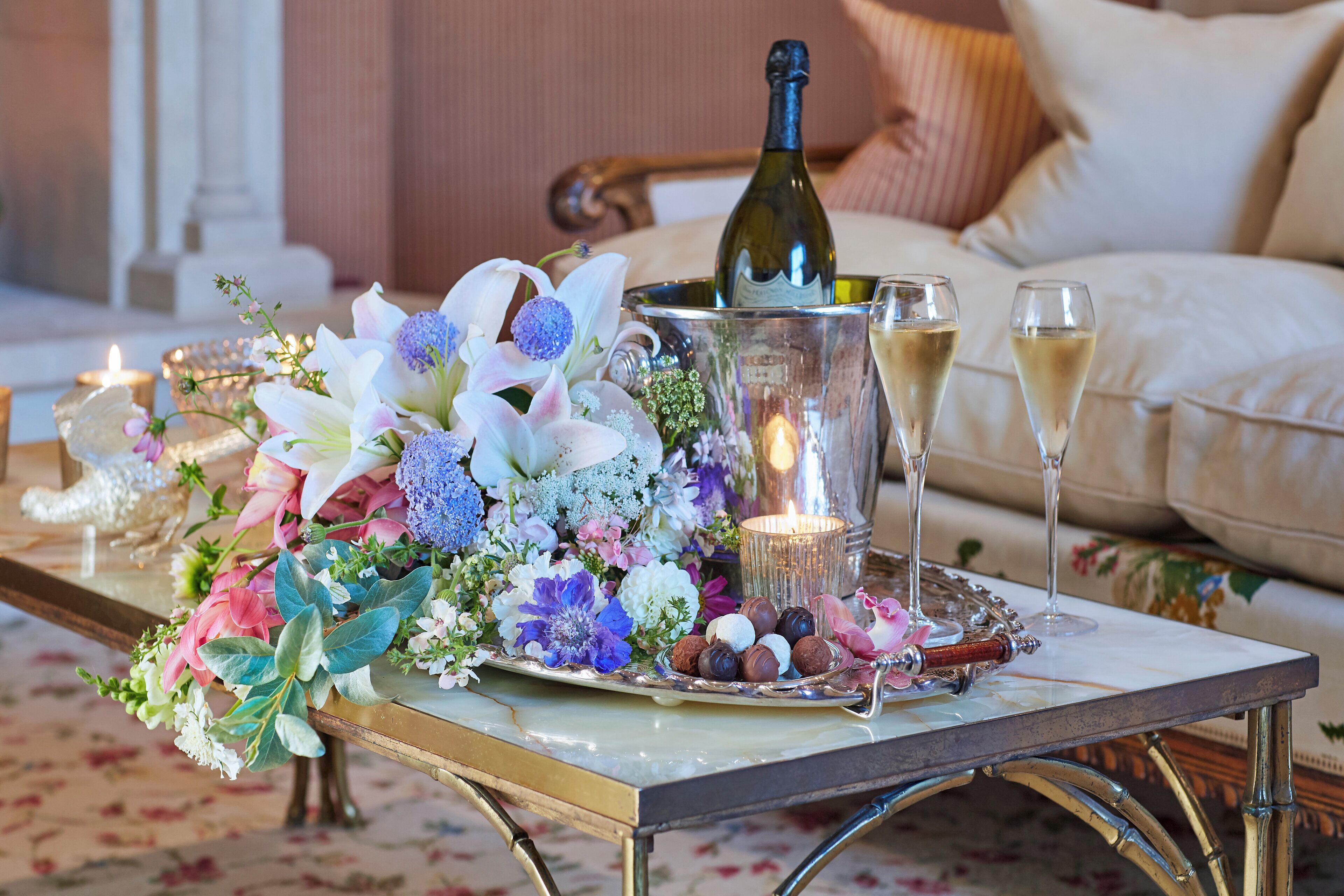 A table with a bottle of champagne, glasses, and a tray of chocolates, surrounded by flowers.