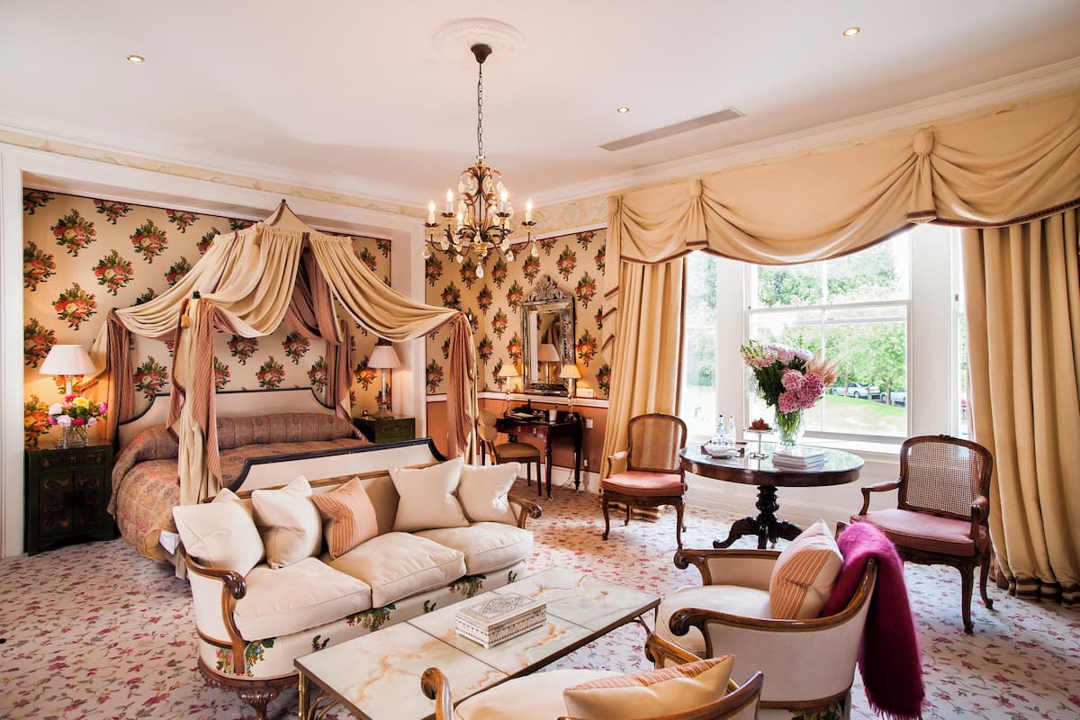 Une chambre à coucher avec un lit à baldaquin, un coin salon avec un canapé et des fauteuils, une table à manger et une grande fenêtre avec des rideaux.