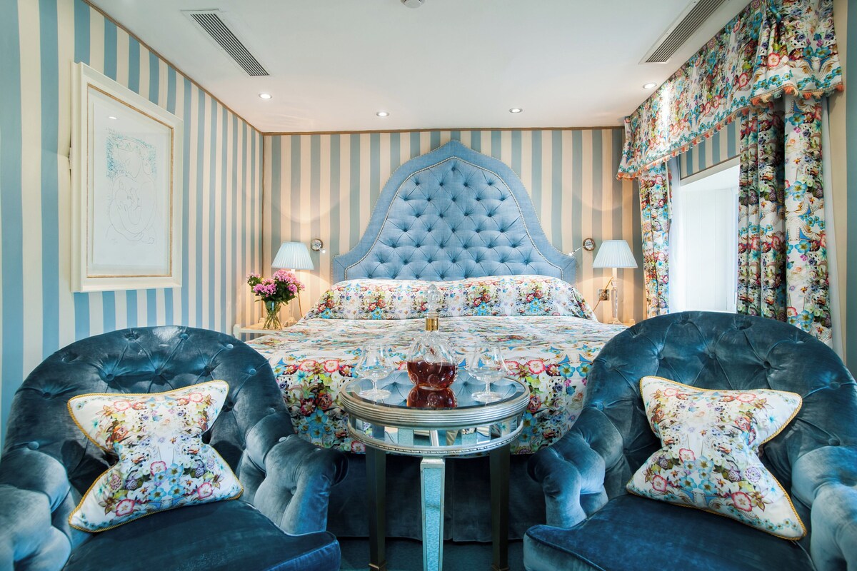 A room with a tufted headboard, floral bedspread, two armchairs with floral cushions, a round table with a glass top, and striped wallpaper.