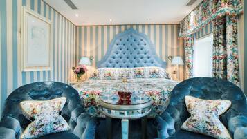 A room with a tufted headboard, floral bedspread, two armchairs with floral cushions, a round table with a glass top, and striped wallpaper.