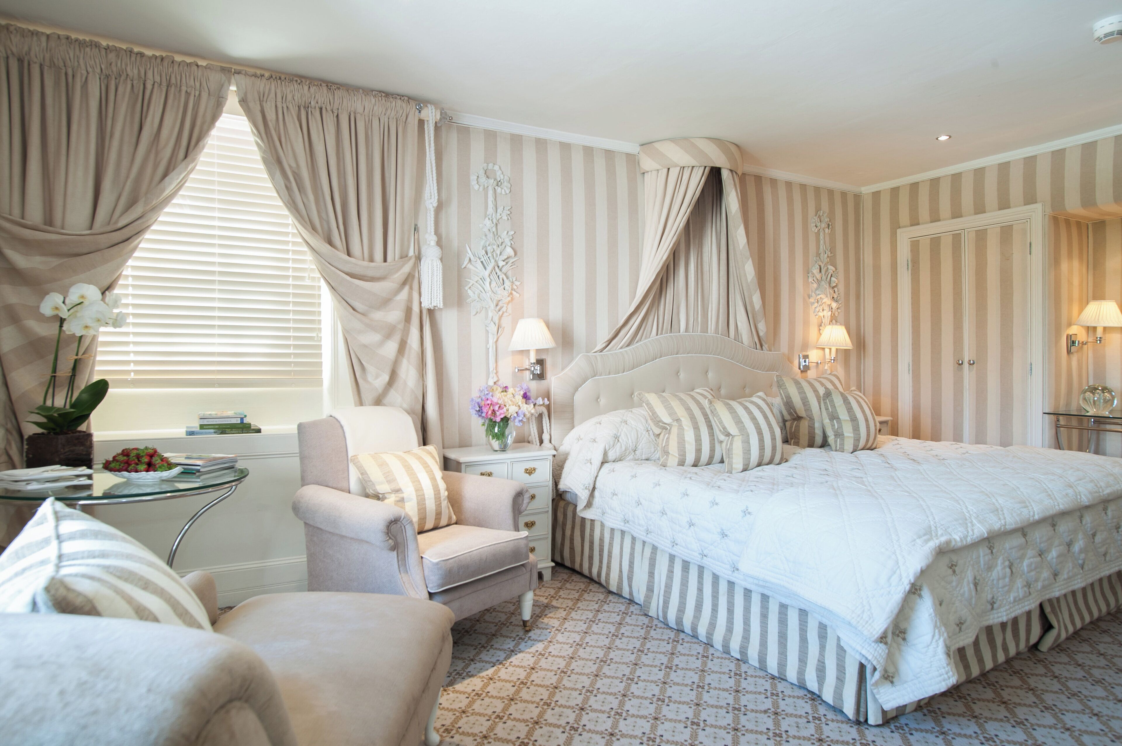 A bedroom with a large bed, a chair, a bedside table with a lamp, and curtains.