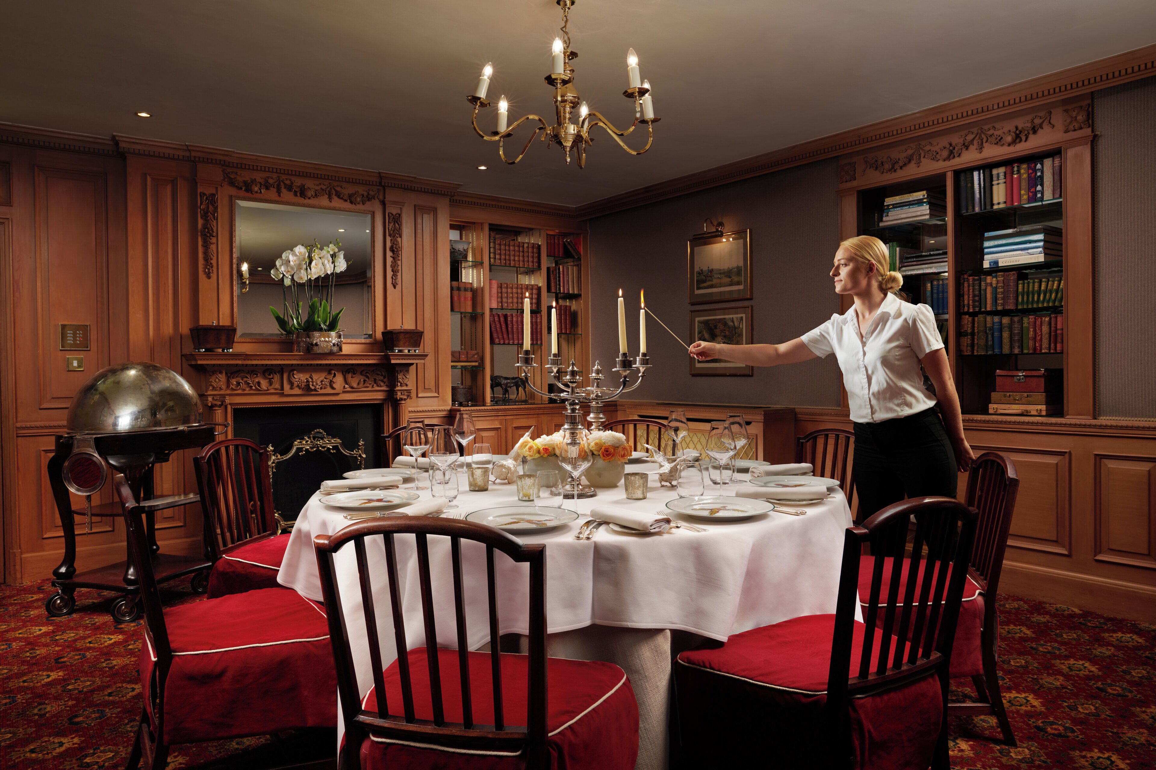 Un comedor formal con candelabro, sillas tapizadas en rojo, una mesa cubierta con mantel blanco y una estantería al fondo.
