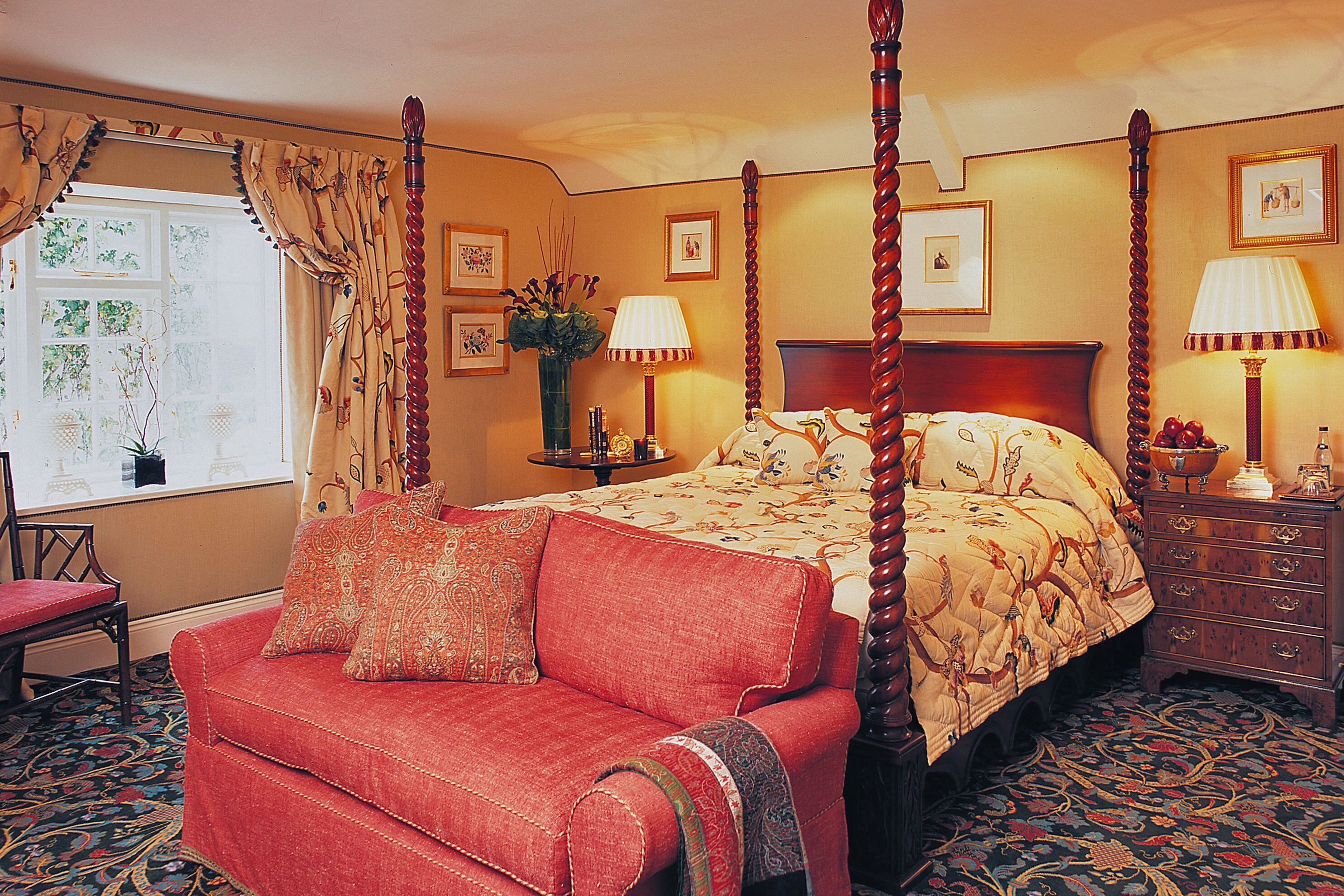 A four-poster bed with a floral bedspread, a red upholstered chair, and a wooden nightstand with a lamp.