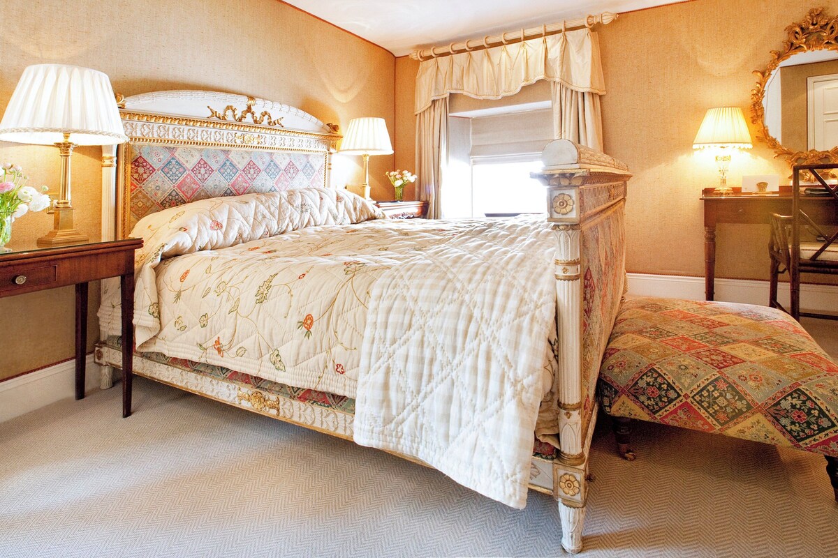 A classic four-poster bed with a quilted bedspread, a nightstand with a lamp, and a chair with a cushion.