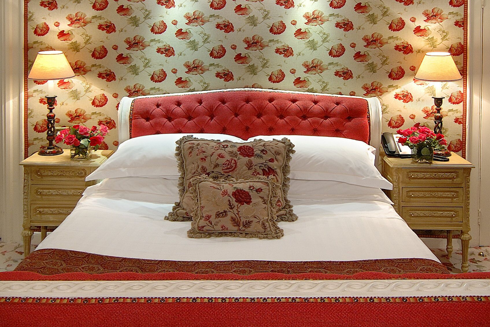 A neatly made bed with floral-patterned pillows and a red upholstered headboard. There are two bedside tables with lamps and a vase of flowers on each.