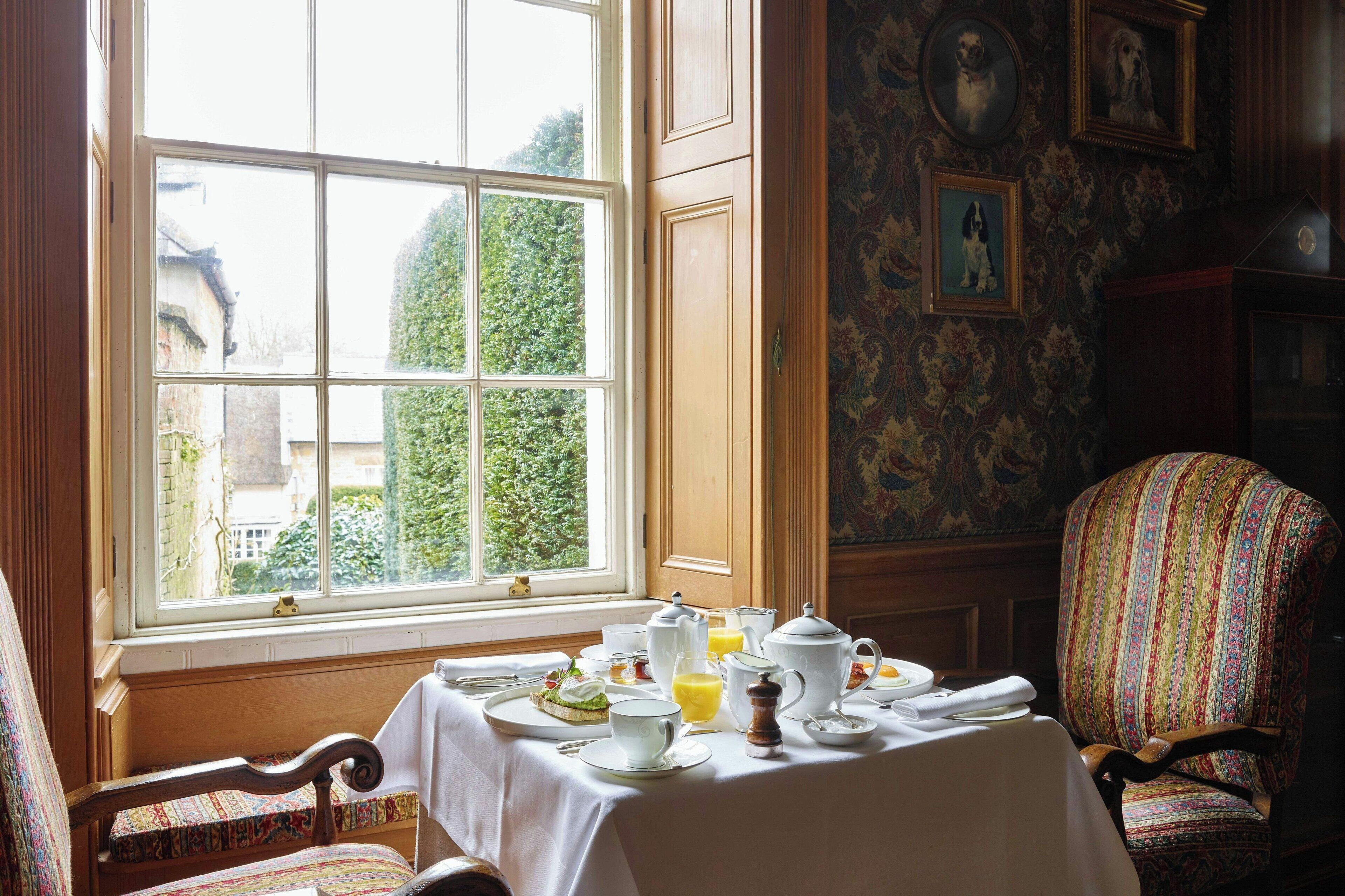 A dining area with a table set for breakfast, a window with a view of greenery, and a patterned wall with framed pictures.