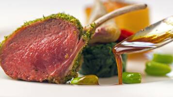 Primer plano de un plato de carne cocida con costra dorada, acompañado de una cuchara y algunas verduras verdes.