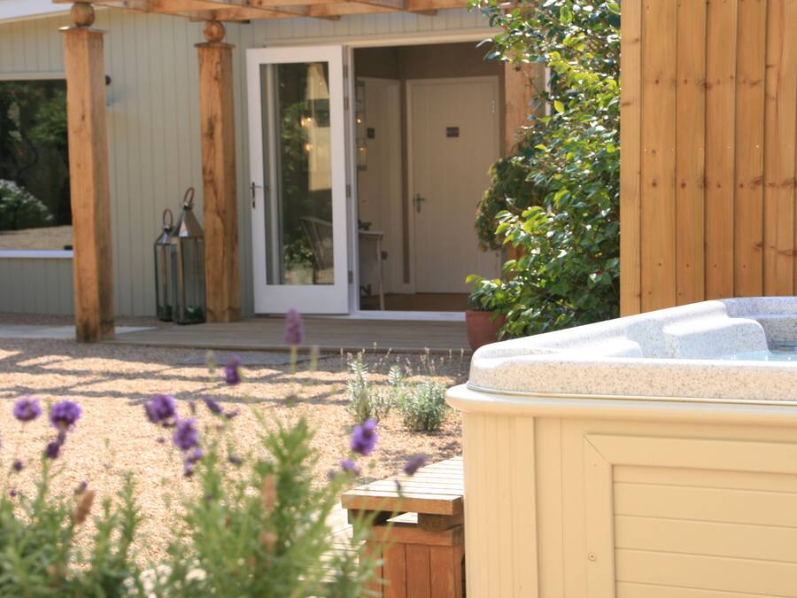 A hot tub in a garden area with a wooden pergola and a door leading to a covered patio.