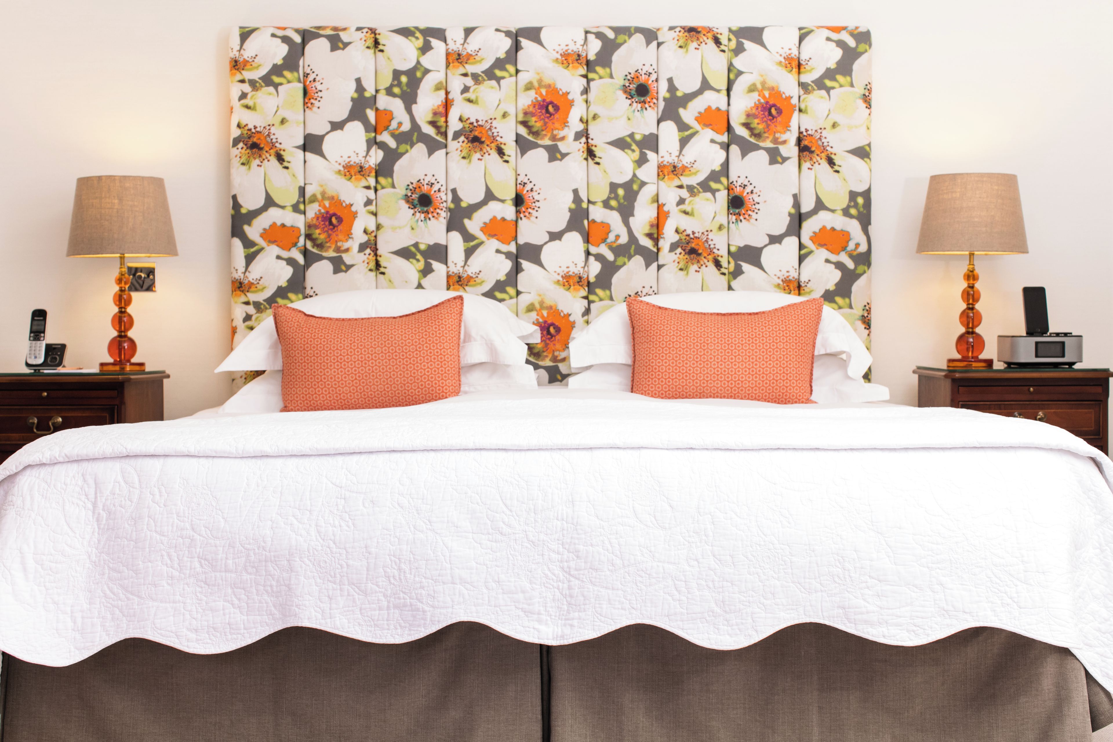 Une chambre d’hôtel avec une grande tête de lit ornée de motifs floraux, deux oreillers, un couvre-lit blanc et deux tables de chevet avec des lampes et une radio.