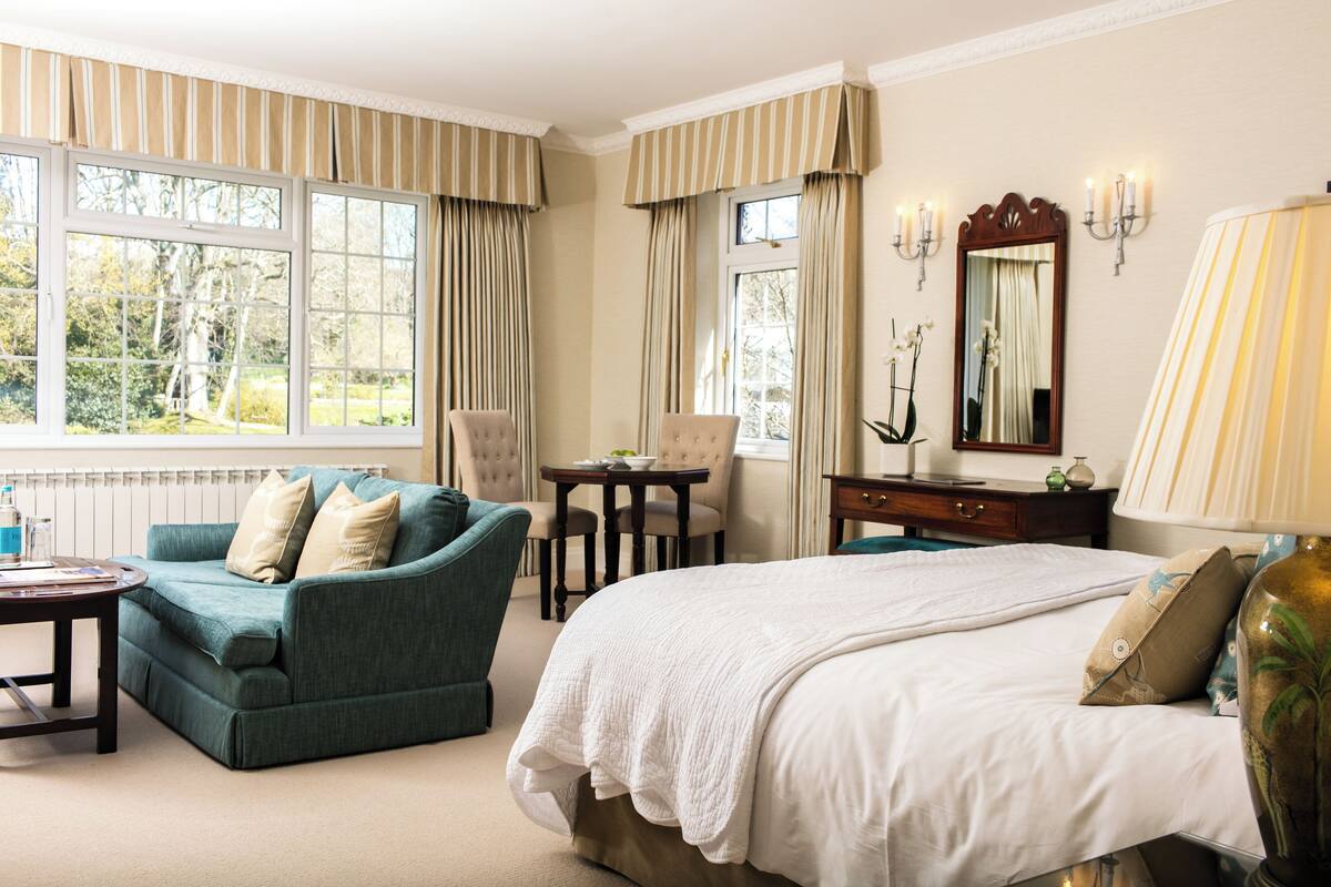 A hotel room with a large bed, a green armchair, a small table, and a desk. There are curtains on the windows, a mirror, and a lamp on the desk.