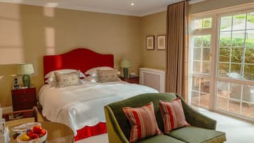 A bedroom with a large bed, a green armchair, a bedside table with a lamp, and a view of the garden through a window.