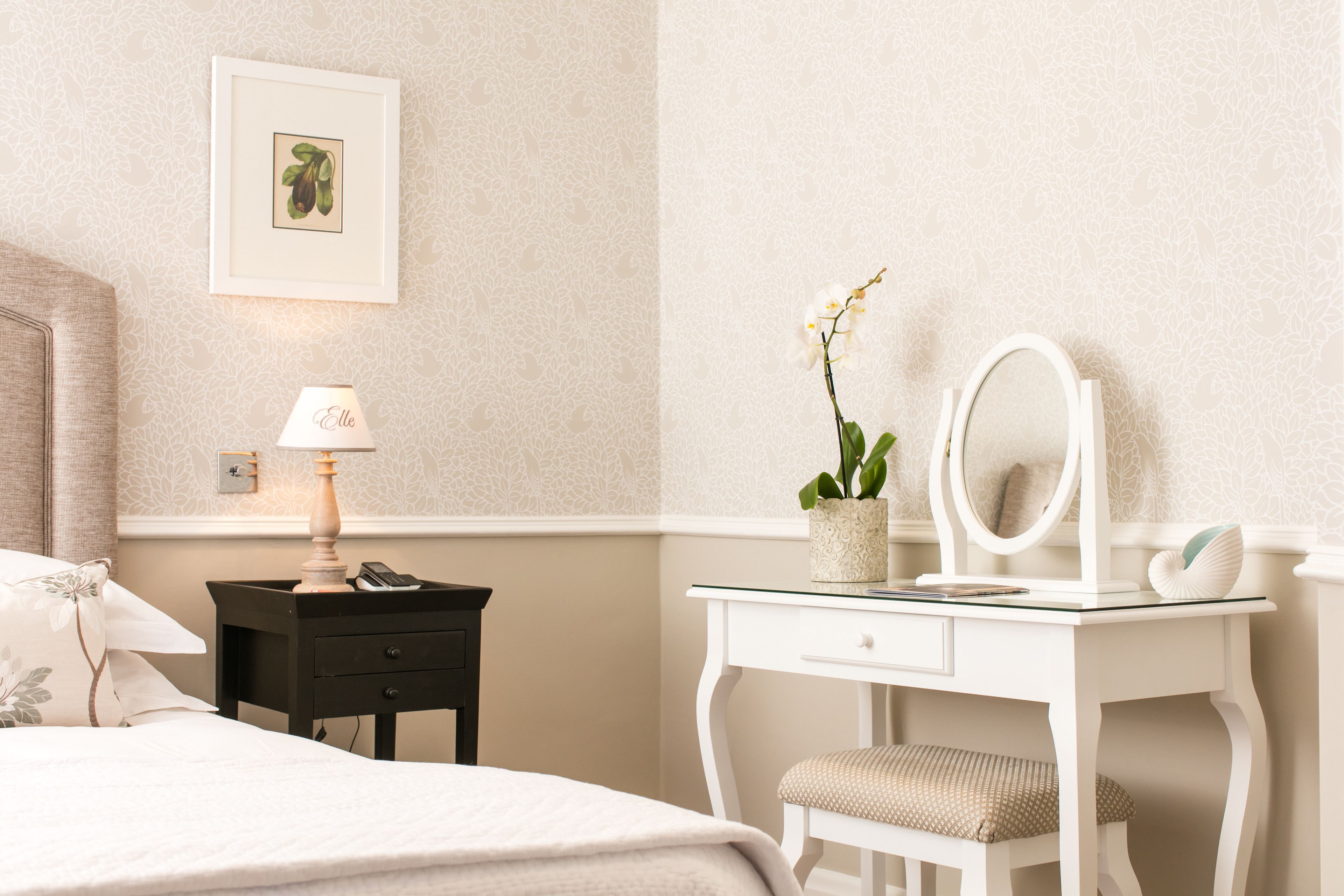 A bedroom with a bed, a nightstand with a lamp, a dresser with a mirror, and a potted orchid.