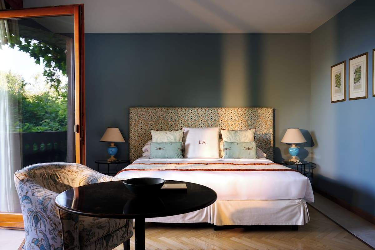 A bedroom with a large bed, two bedside tables with lamps, a chair, and a view of greenery through a sliding door.
