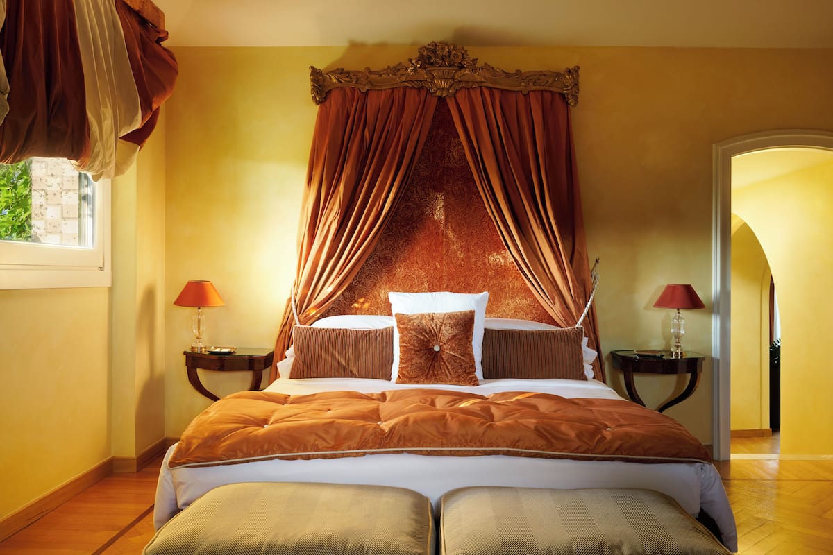 A bedroom with a canopy bed, two bedside tables with lamps, and a view of the outside through a window.