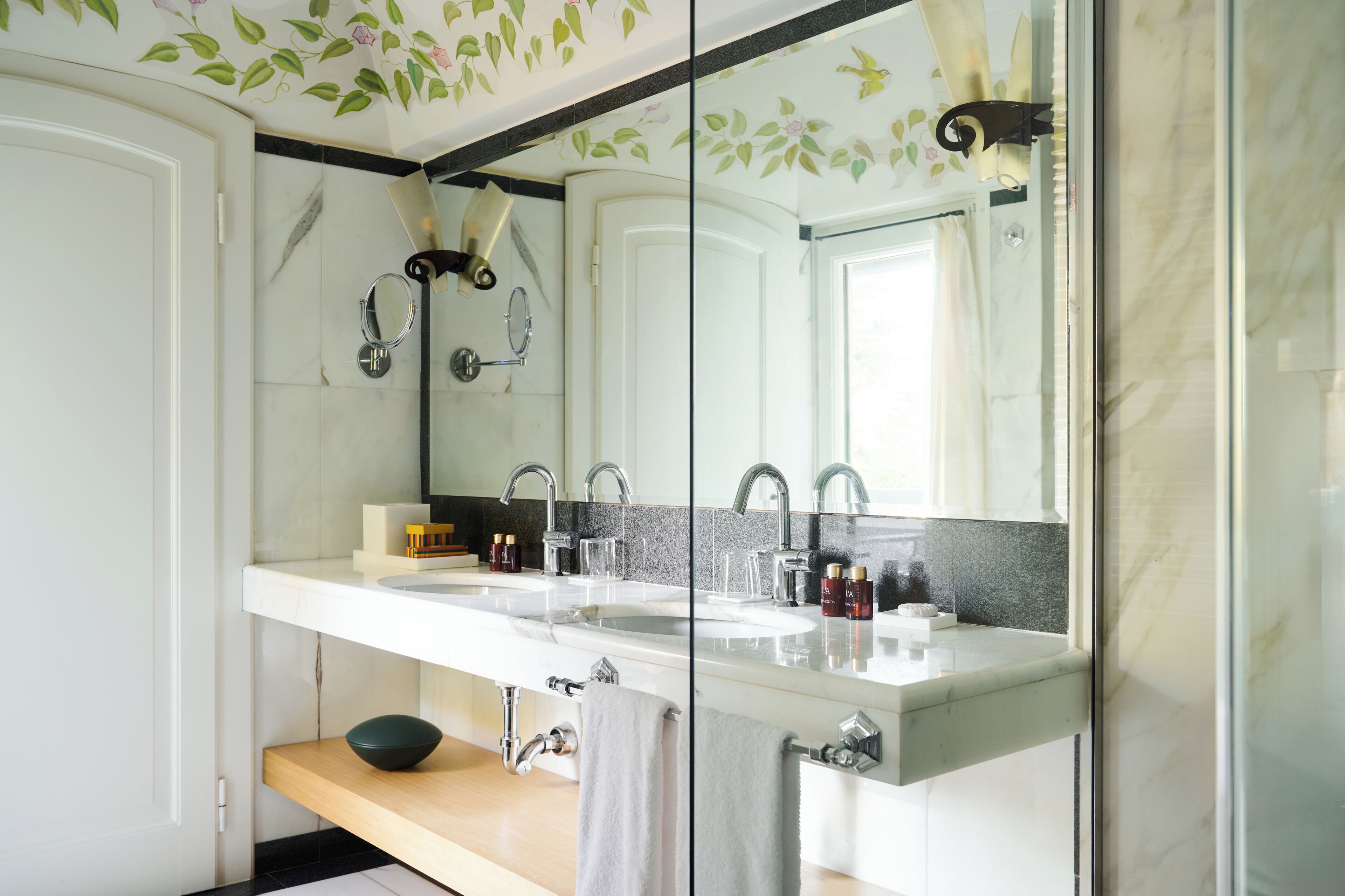 A modern bathroom with a large mirror, two sinks, and a wooden shelf with a bowl.