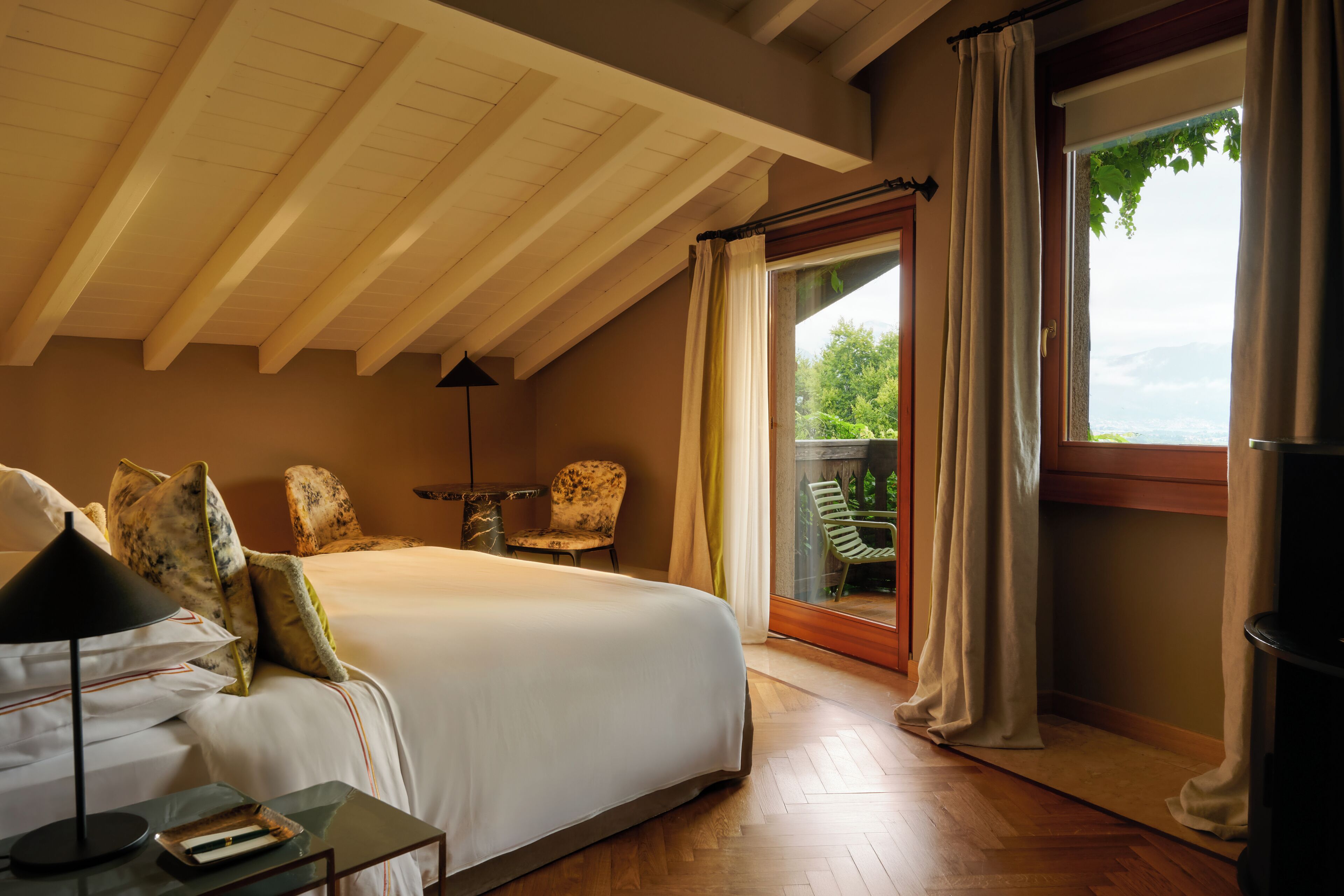 A bedroom with a large bed, a nightstand, a lamp, a chair, and a view of greenery through a window.
