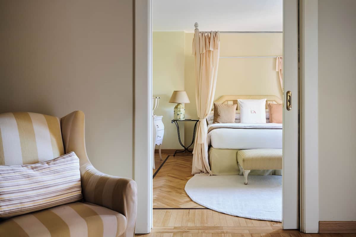 A hotel room with a bed, a bedside table with a lamp, a chair, and a view into another room.