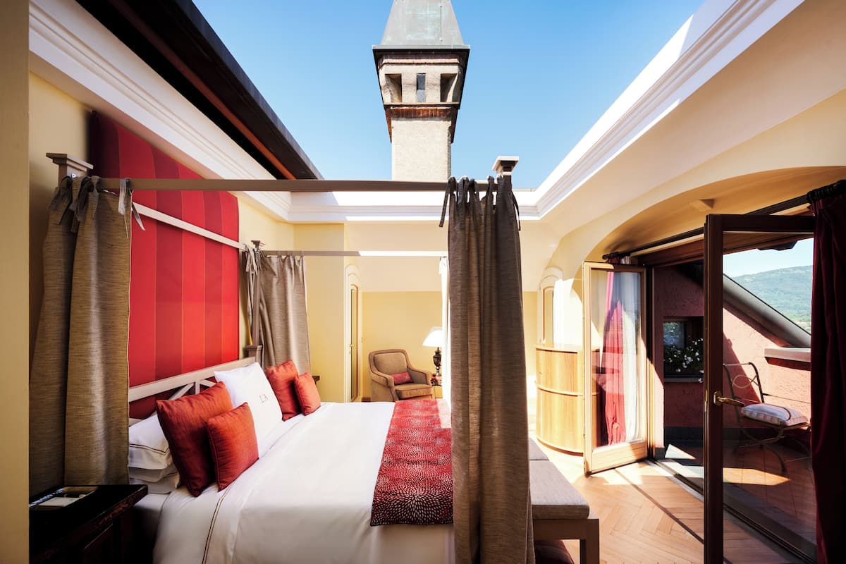 A hotel room with a large bed, red curtains, a view of the sky, and a balcony with a chair.