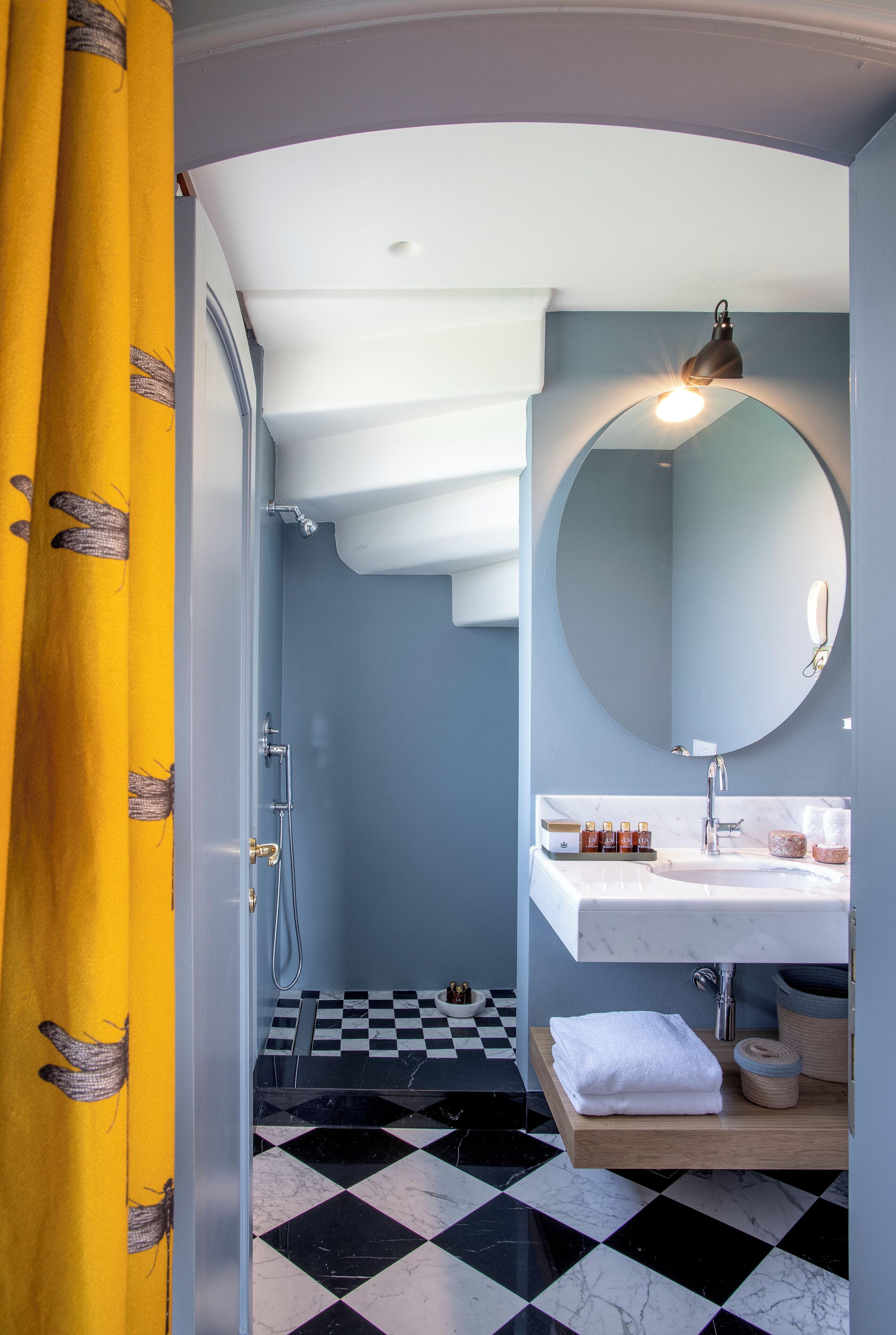 Une salle de bain avec un carrelage à motifs géométriques, un miroir rond, un lavabo blanc et un rideau jaune.