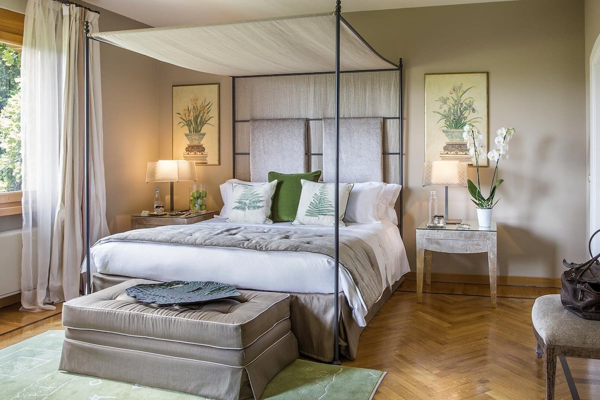 A four-poster canopy bed with a wooden headboard, a nightstand with a lamp, a small table with a lamp, a bench, and a chair.