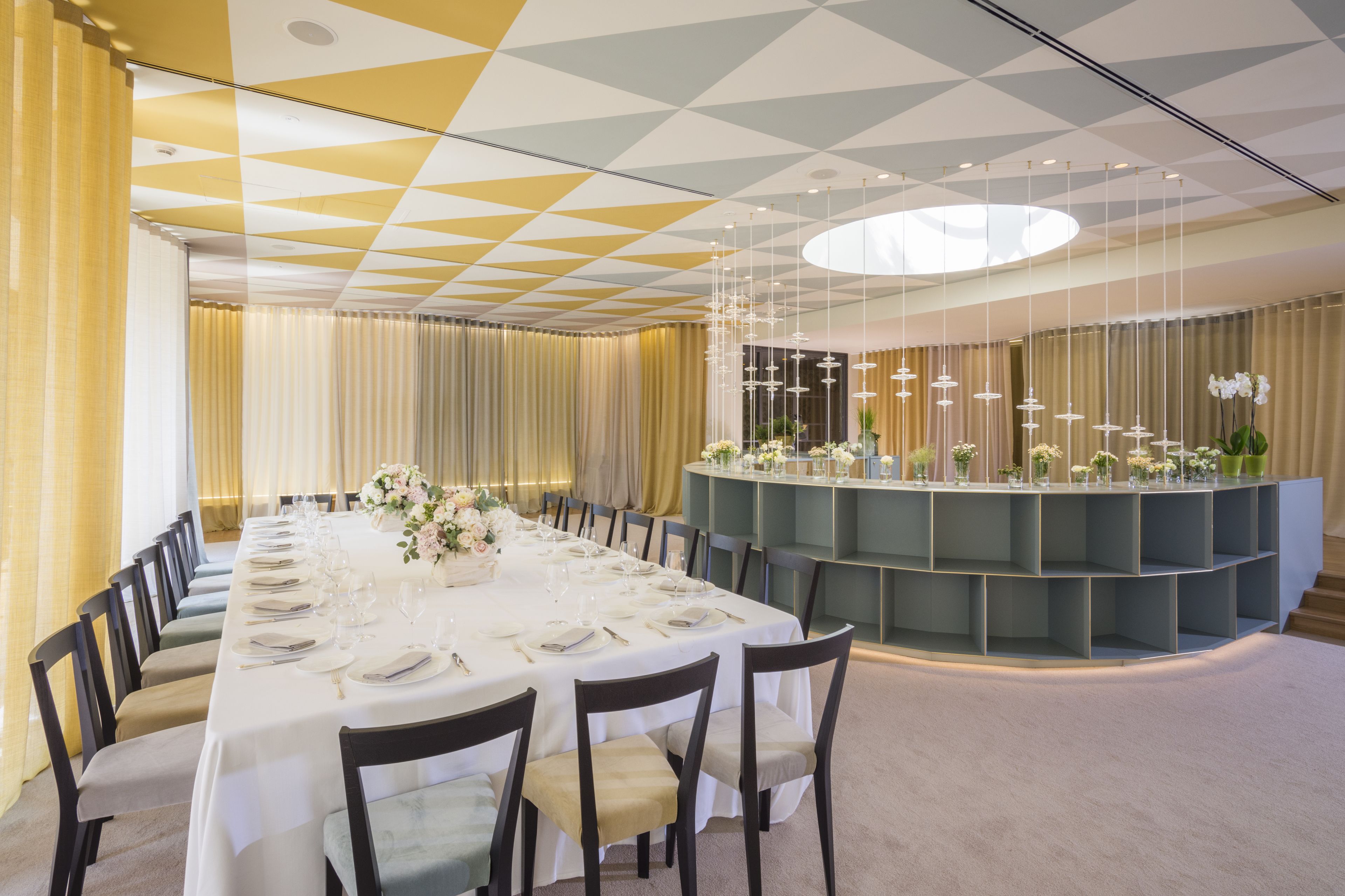 A long dining table set for a formal event, surrounded by chairs, with a decorative centerpiece and a modern, geometric ceiling design.