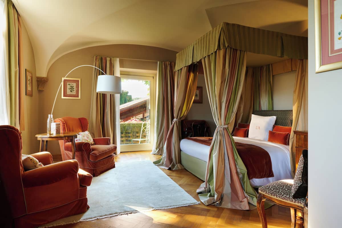A bedroom with a canopy bed, a sitting area with a chair and a table, and a view of the outside.