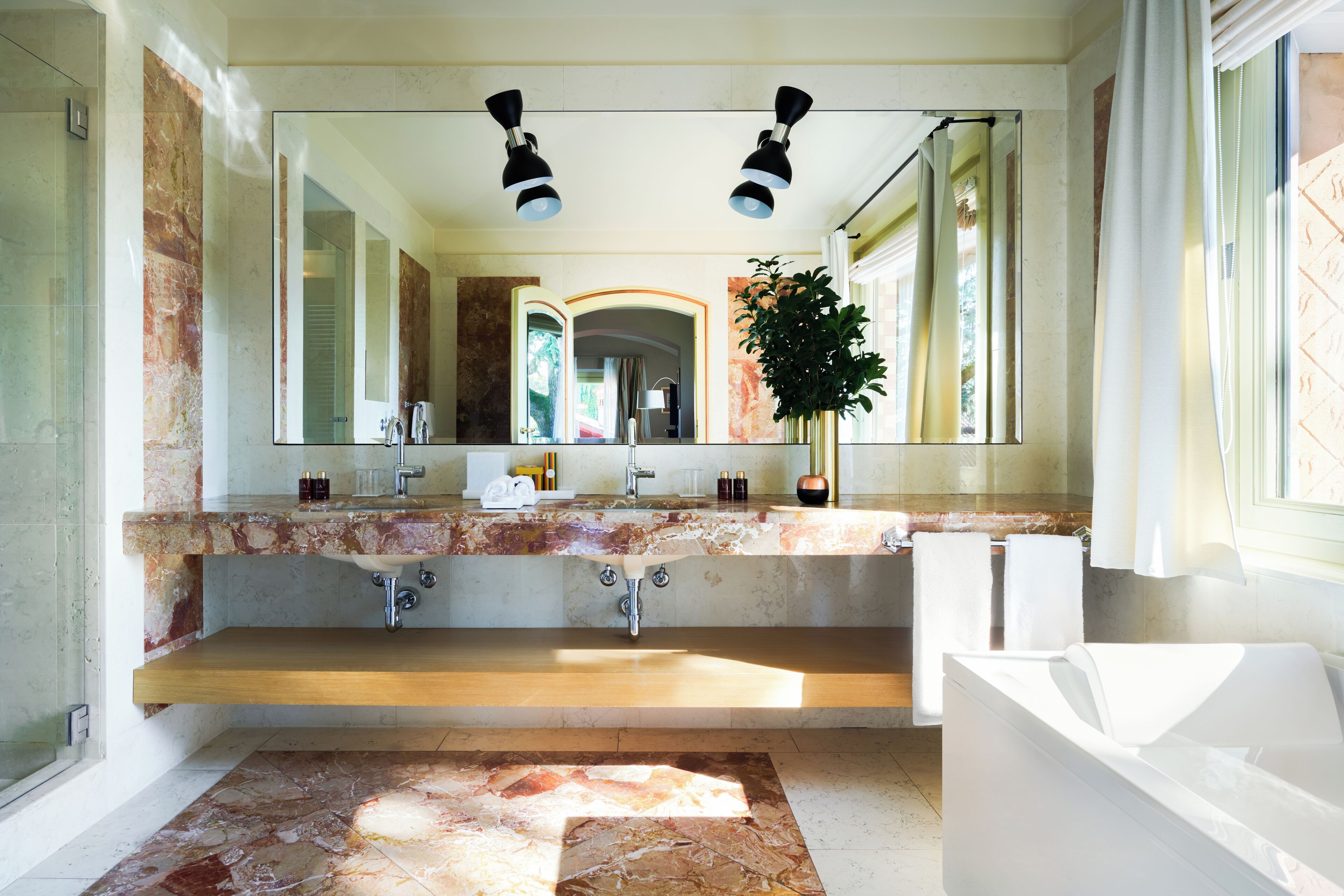A modern bathroom with a large mirror, two sinks, a bathtub, and a potted plant.