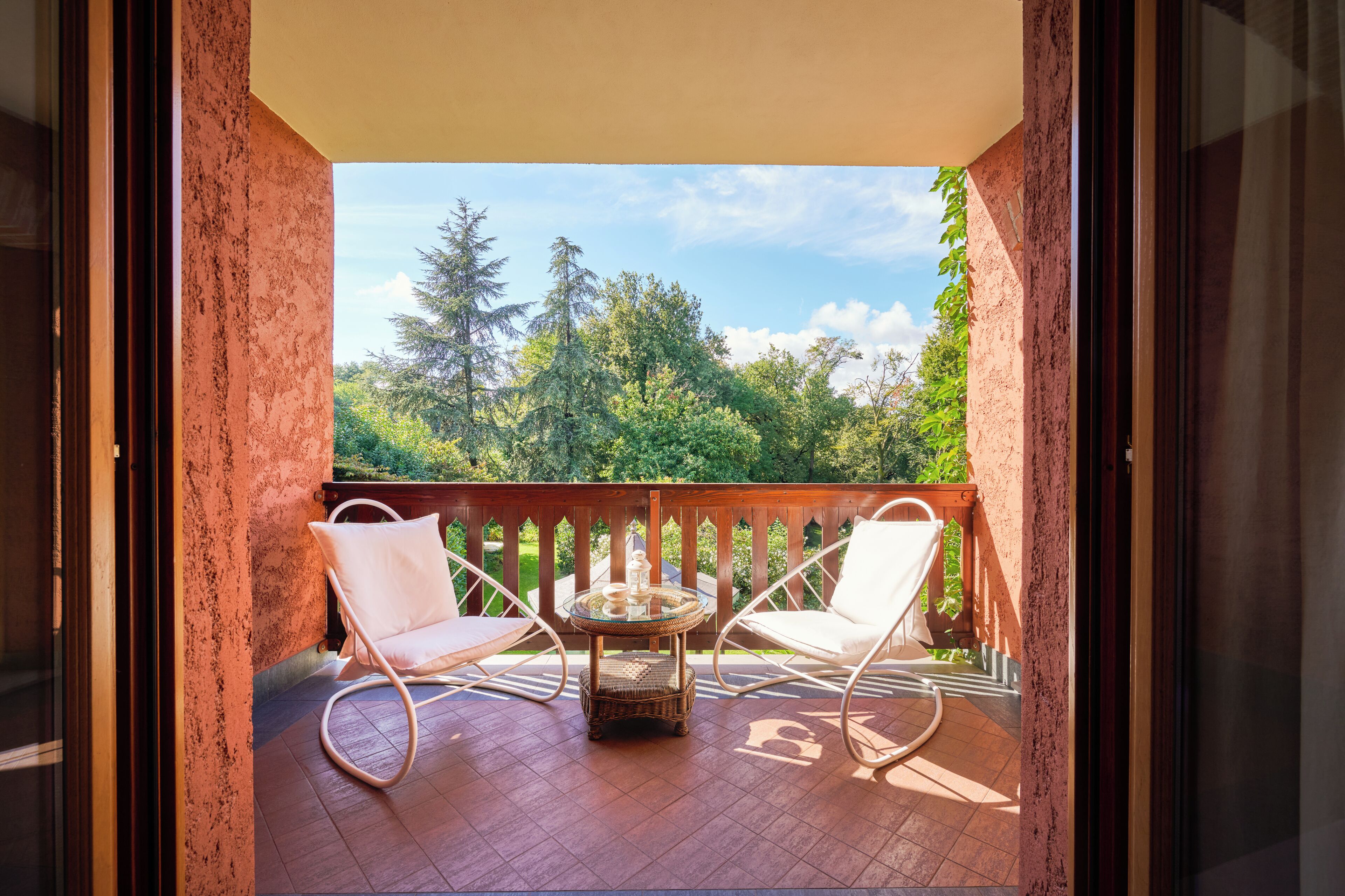 A balcony with two white wicker chairs and a glass table, overlooking a forested area.