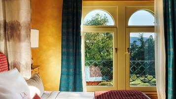 Quarto de hotel com cama, mesa de cabeceira, luminária e vista para uma varanda com vegetação.