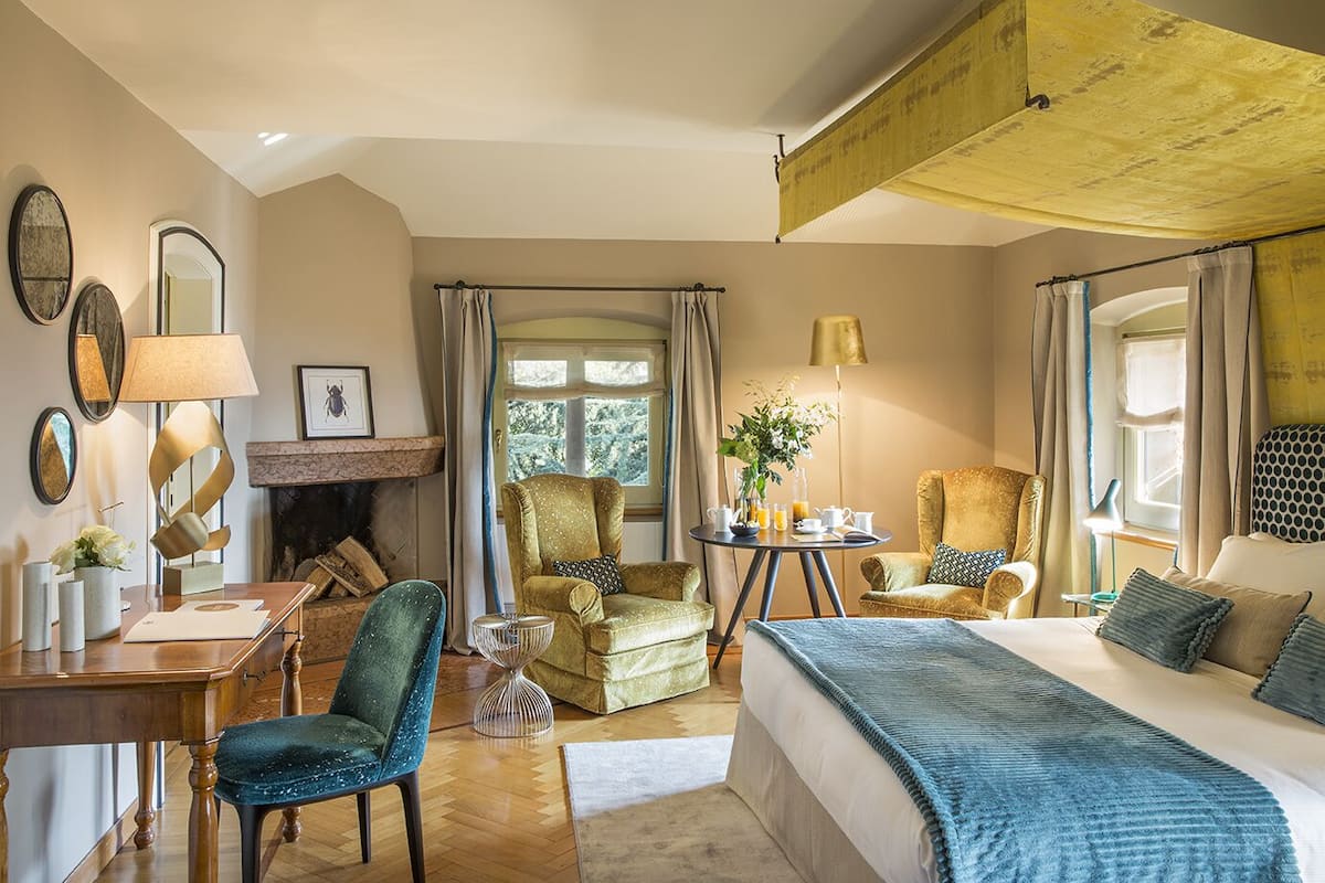A cozy hotel room with a bed, a desk, a chair, a fireplace, and a dining table with a vase of flowers.