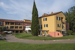 A multi-story building with a yellow facade and green shutters, surrounded by a well-maintained lawn and a tall cypress tree. - La Meridiana Relais & Chateaux (Garlenda)