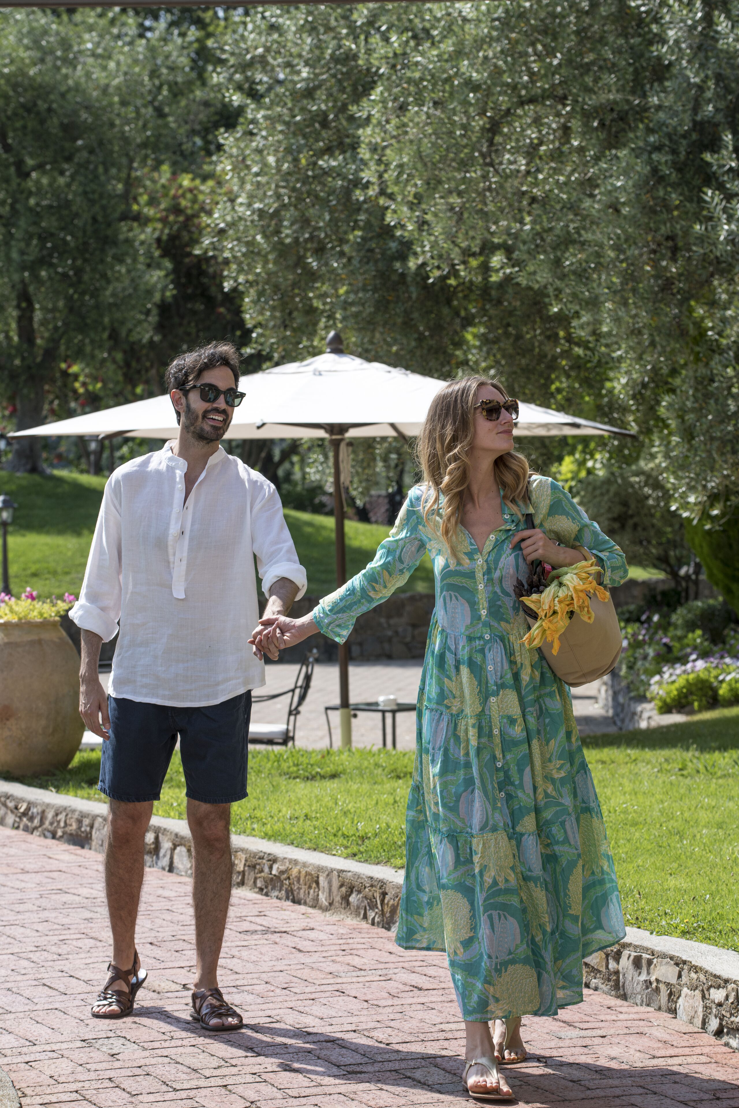 A couple walking hand in hand on a paved path in a park-like setting with greenery and a large umbrella in the background.
