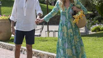A couple walking hand in hand on a paved path in a park-like setting with greenery and a large umbrella in the background.