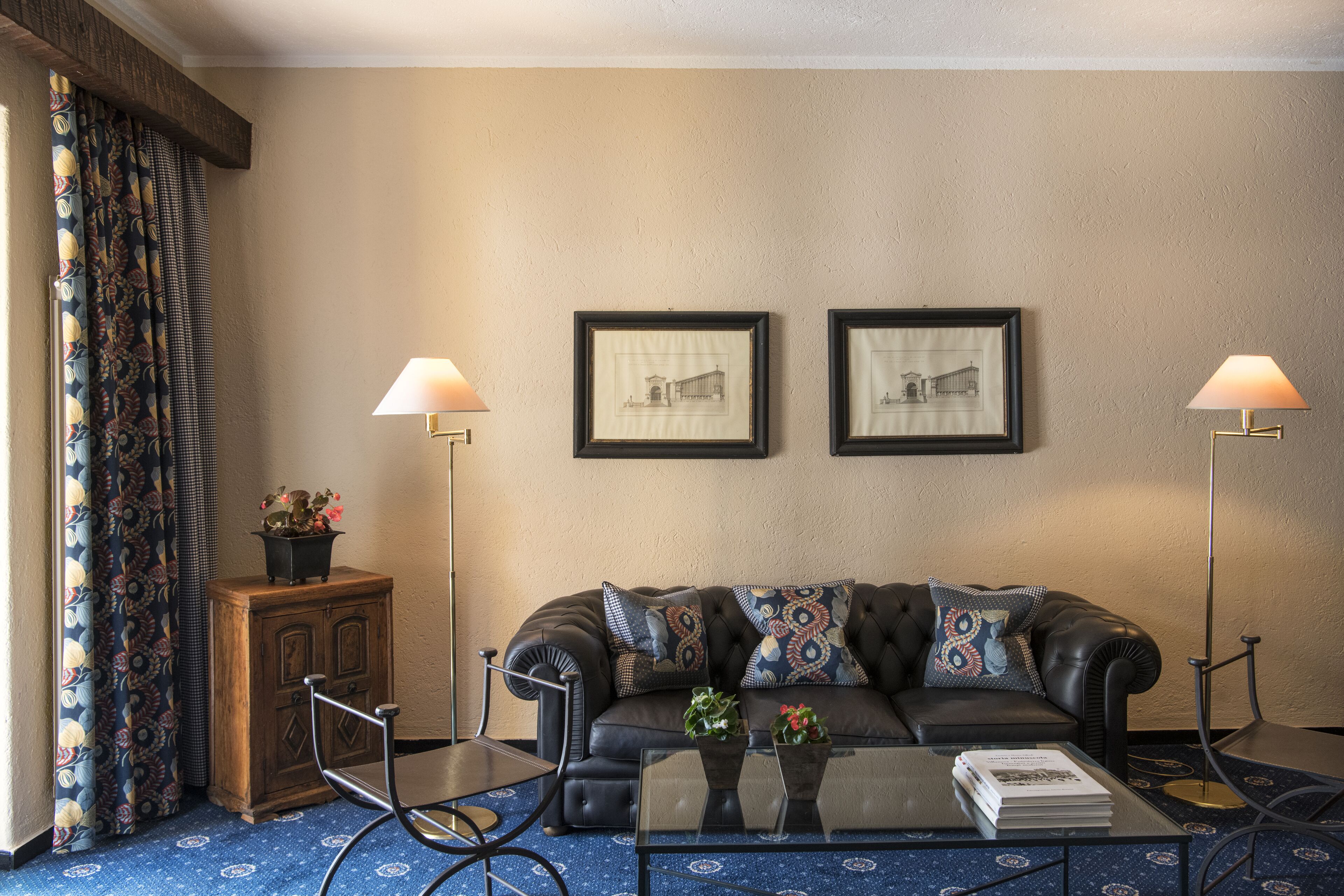Ein Wohnzimmer mit einer Ledersofa, zwei gerahmten Bildern an der Wand, einem Glastisch mit Büchern, einer kleinen Holzkommode und zwei Stehlampen.