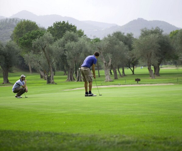 A golf course with two players, one standing and one crouching, with trees and mountains in the background.
