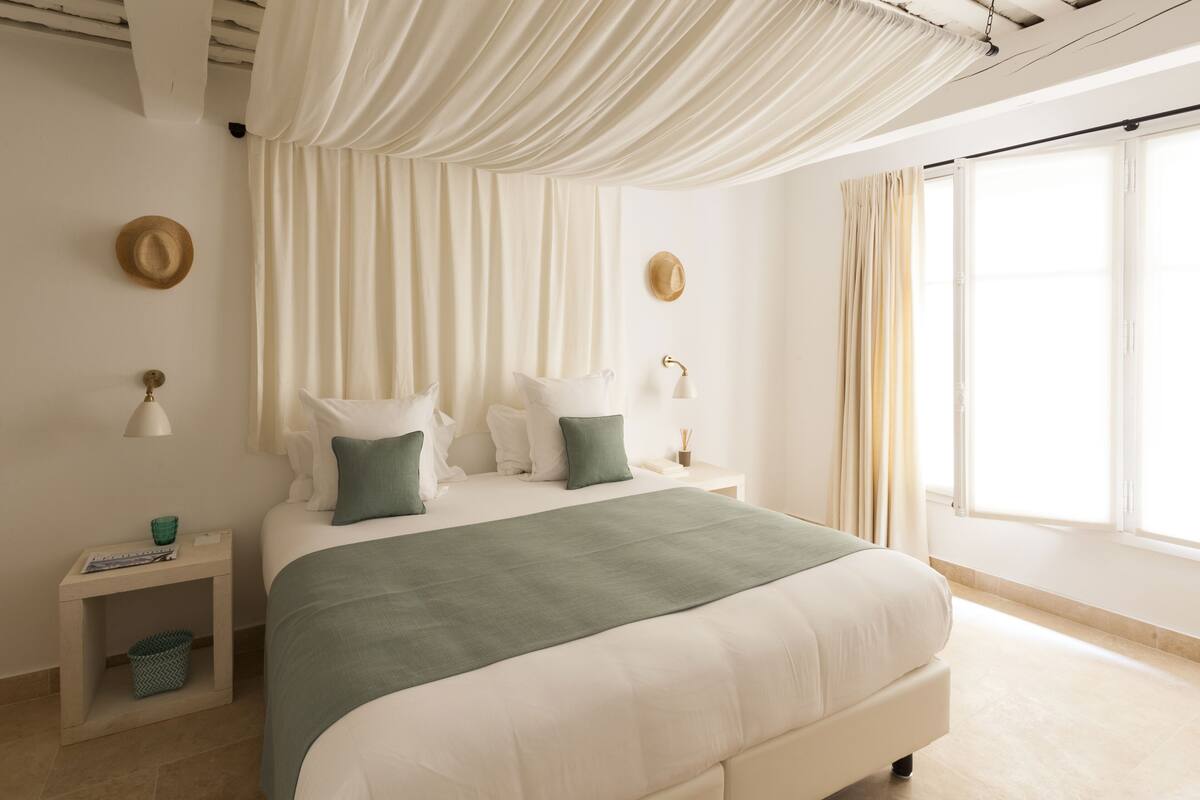 Ein Schlafzimmer mit einem großen Bett, einem Nachttisch, einer Lampe und einem Fenster mit Vorhängen.