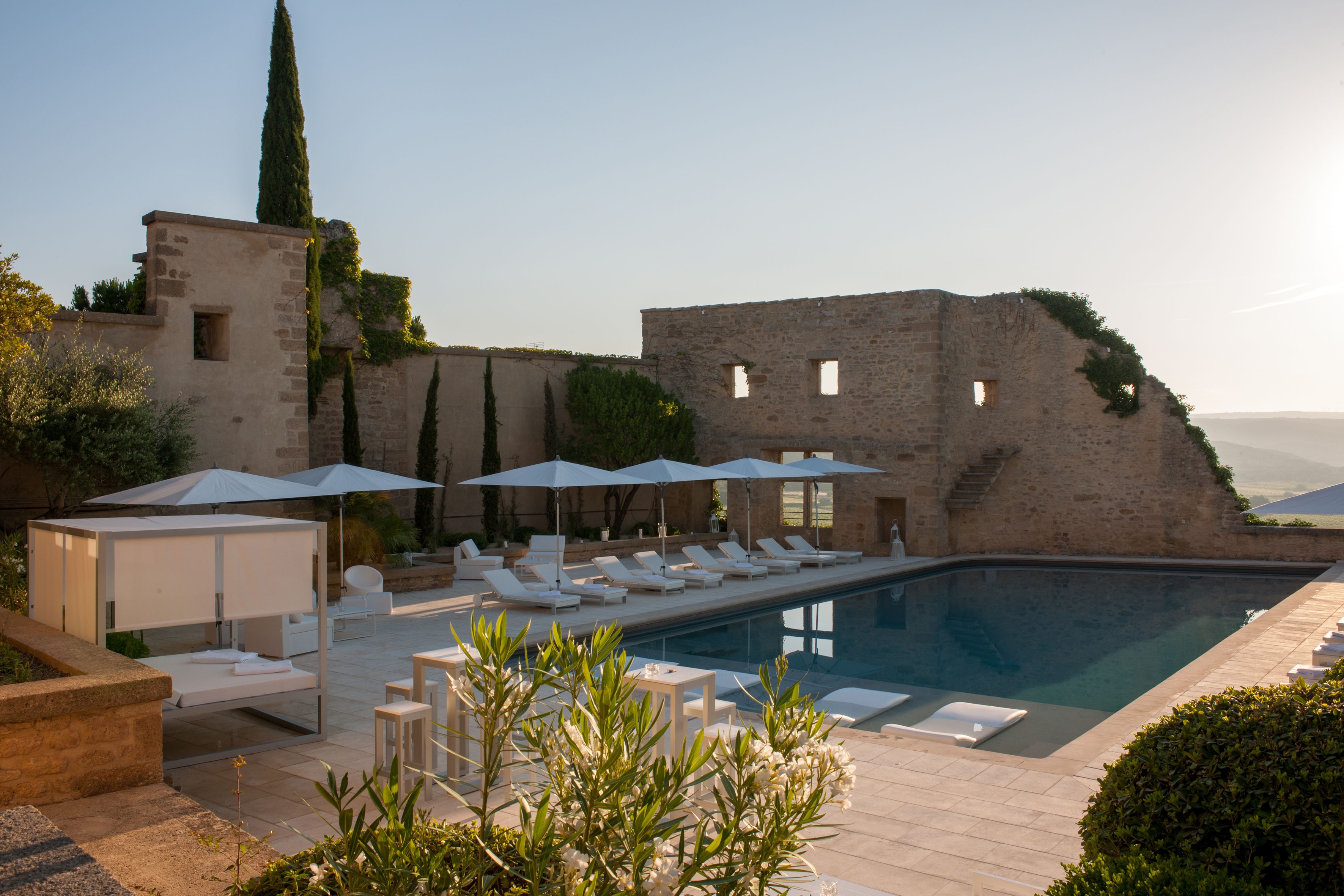 A pool area with lounge chairs and umbrellas, a stone wall, and a tall tree.