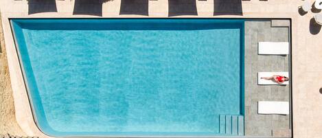 Vista aérea de una piscina rectangular con áreas de descanso adyacentes, que incluyen tumbonas blancas y sombrillas.