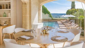 Comedor con vista a la piscina, con una mesa redonda para cuatro personas, sillas blancas y un arco de piedra.