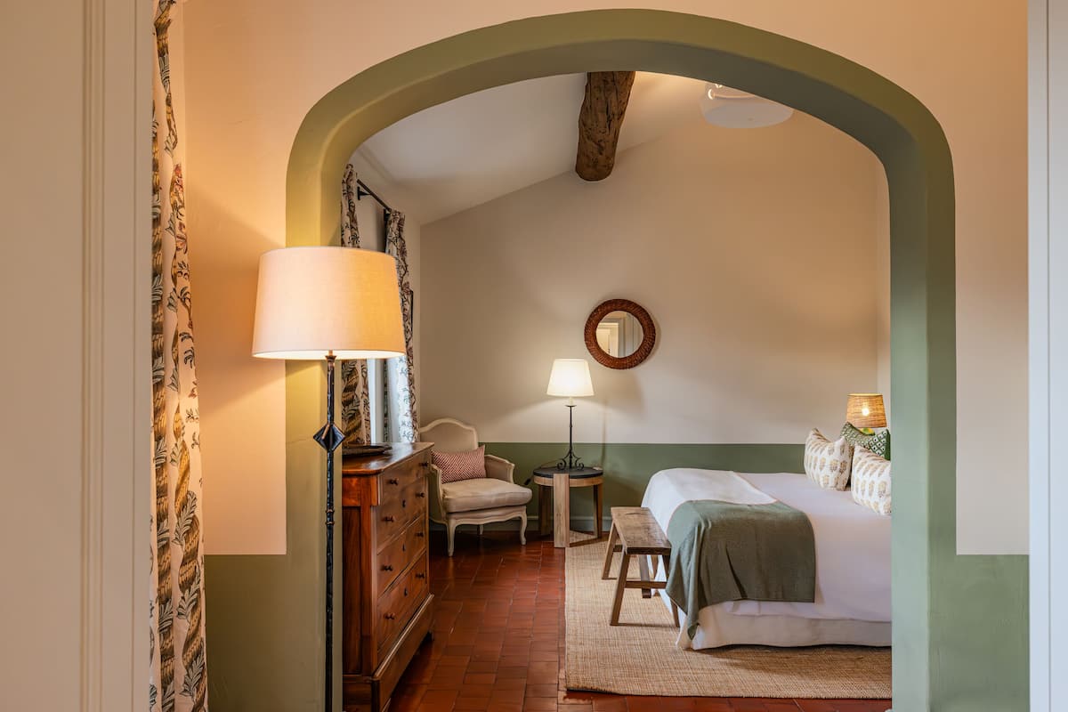 Een knus slaapkamer met een houten dressoir, een nachtkastje met een lamp, een bed met een groene deken en een spiegel aan de muur.