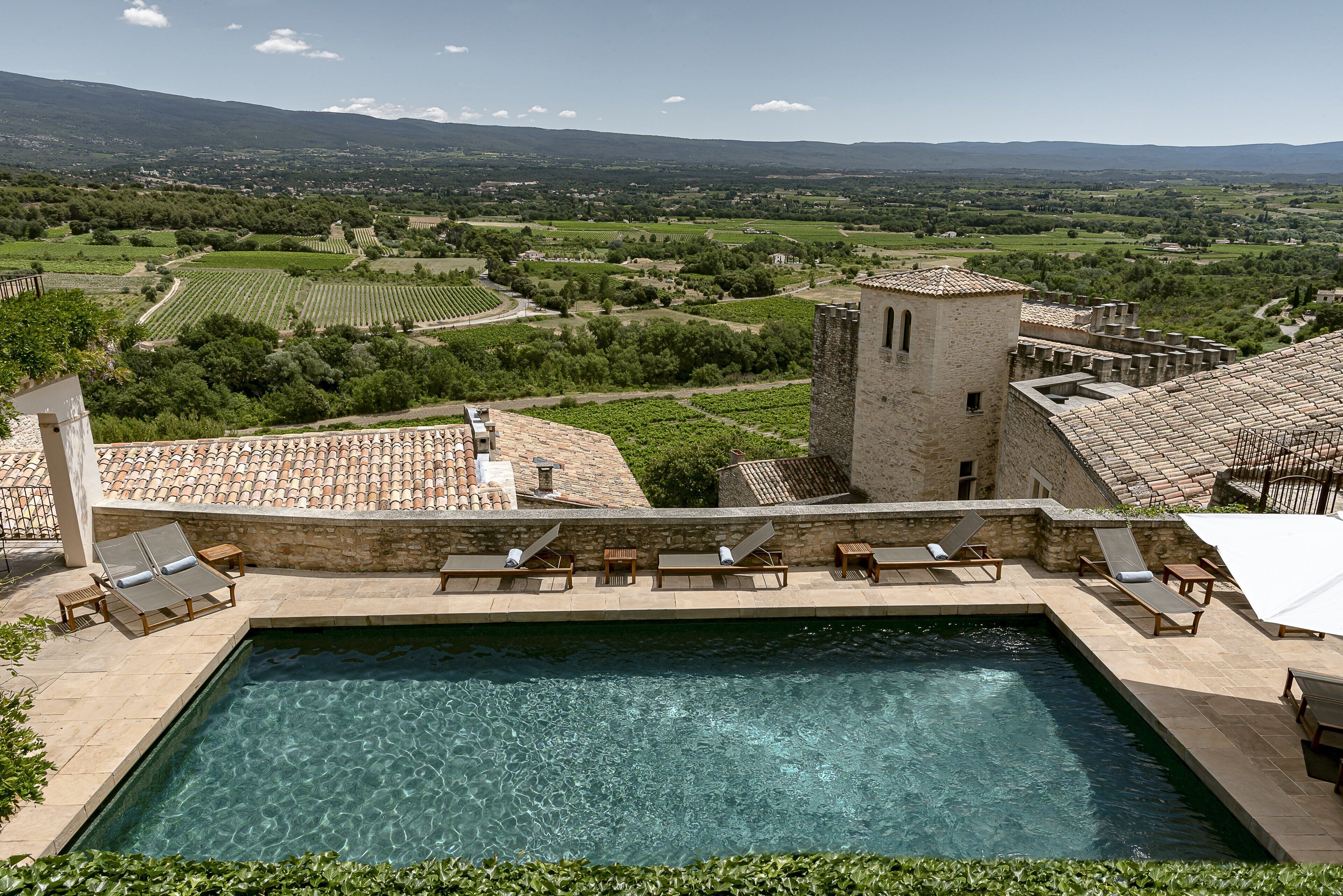 Un área de piscina con agua cristalina, rodeada de sillas de descanso y un edificio de piedra con una torre.