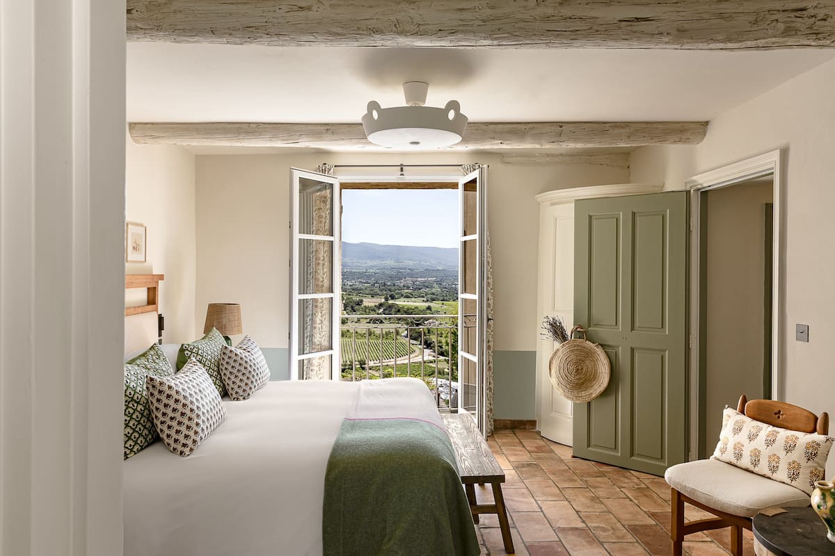 Een slaapkamer met een groot bed, uitzicht door een geopende deur, een groene deur, en een stoel met een kussen.
