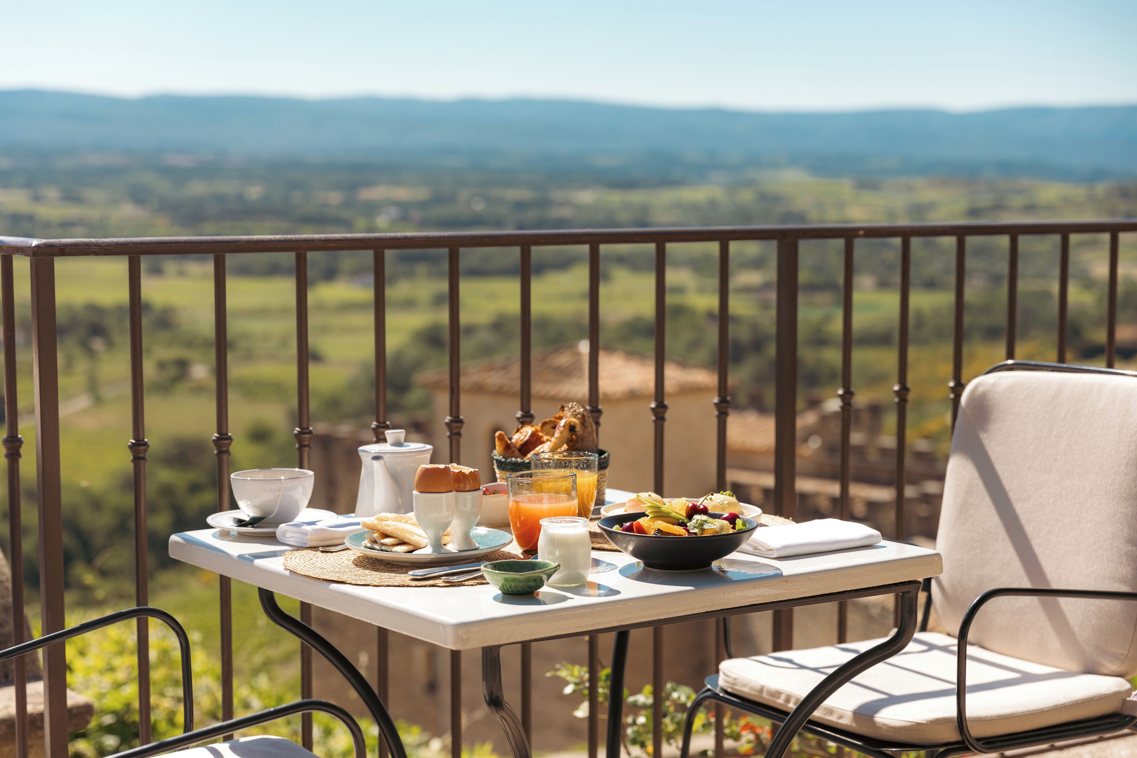 Un balcon avec une table préparée pour le petit-déjeuner, comprenant des œufs, des toasts et un panier de fruits, donnant sur un paysage magnifique.