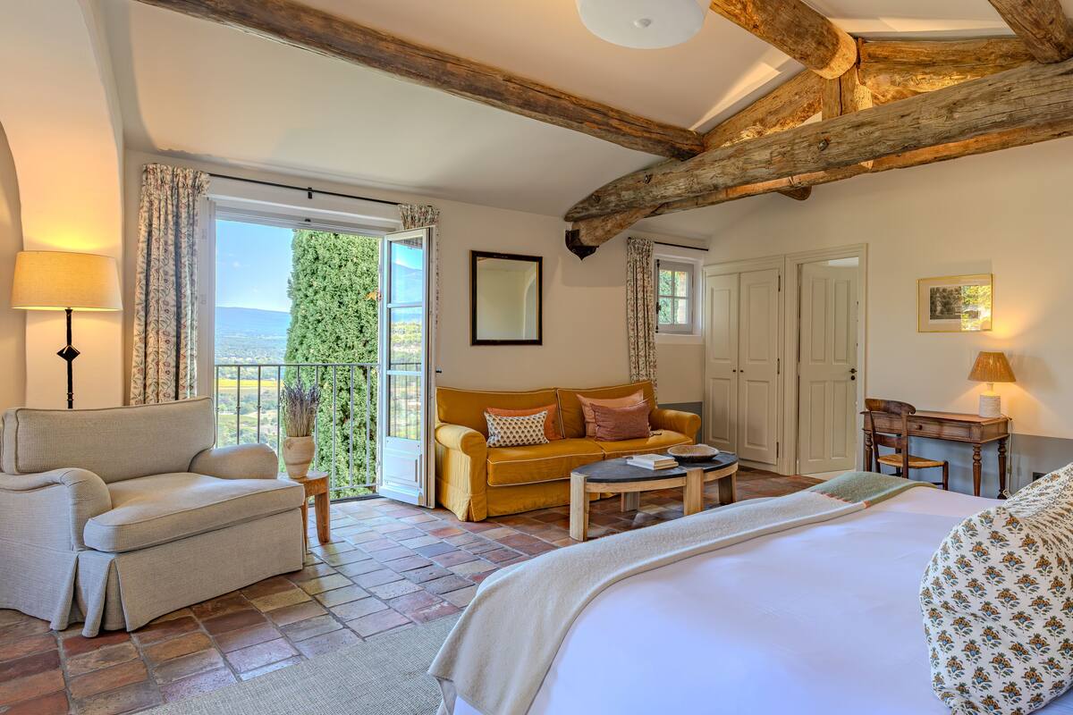 Een gezellige kamer met een bed, een bank, een stoel en een tafel. Er is een balkon met uitzicht op de omgeving.