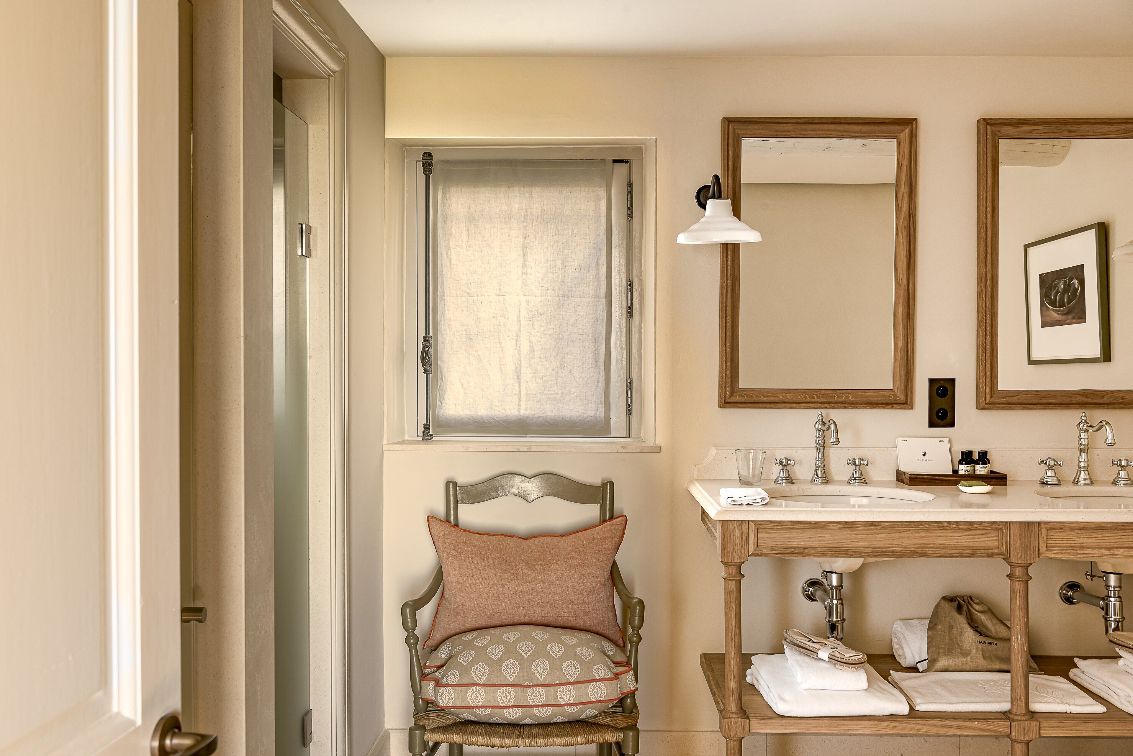 Une salle de bain avec un meuble-lavabo en bois, une chaise avec un coussin, un miroir et une fenêtre avec un rideau.