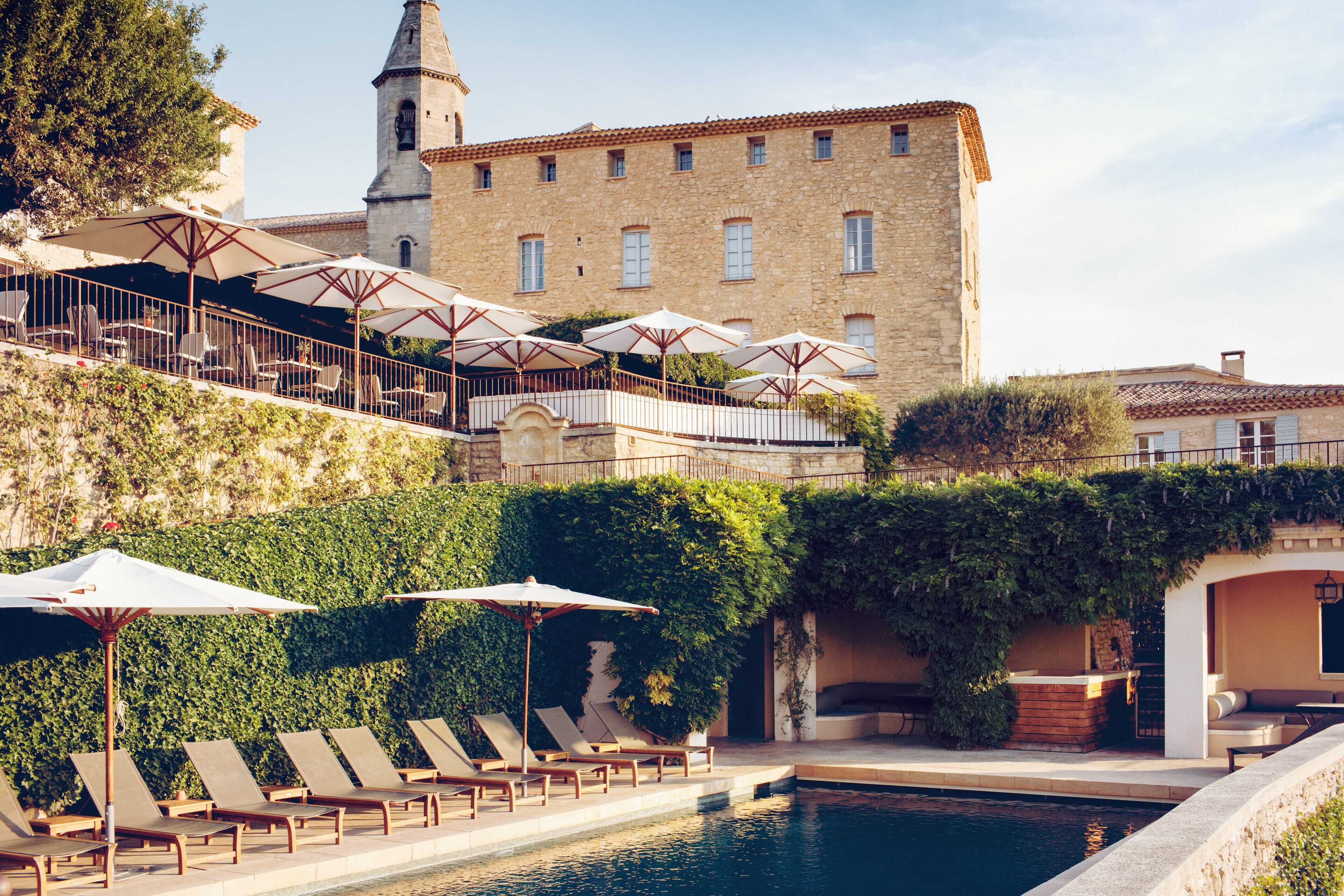 A poolside area with lounge chairs and umbrellas, a stone building with a tower, and a lush green wall.